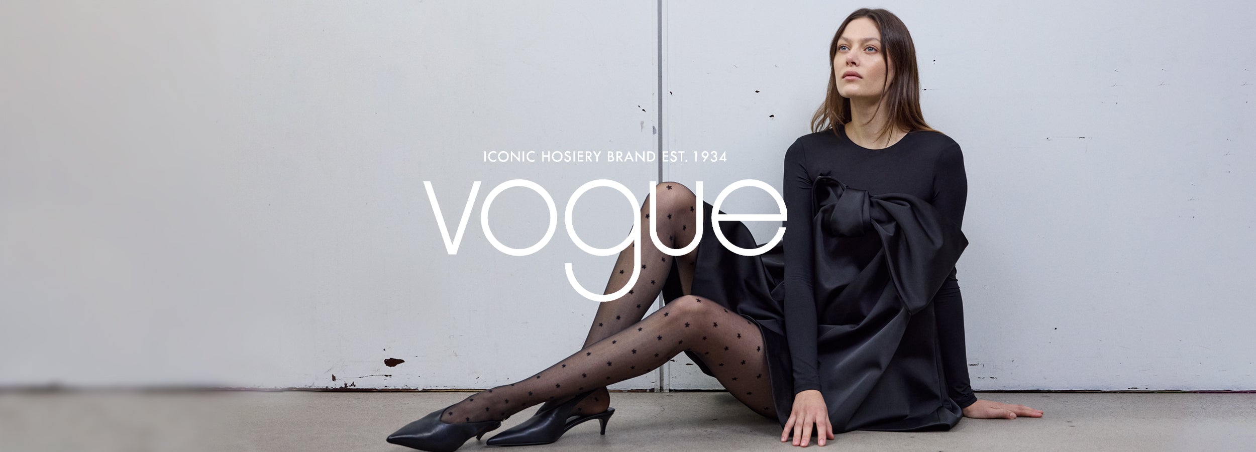 Woman in a black dress and Vogue star patterned hosiery sitting against a white wall.
