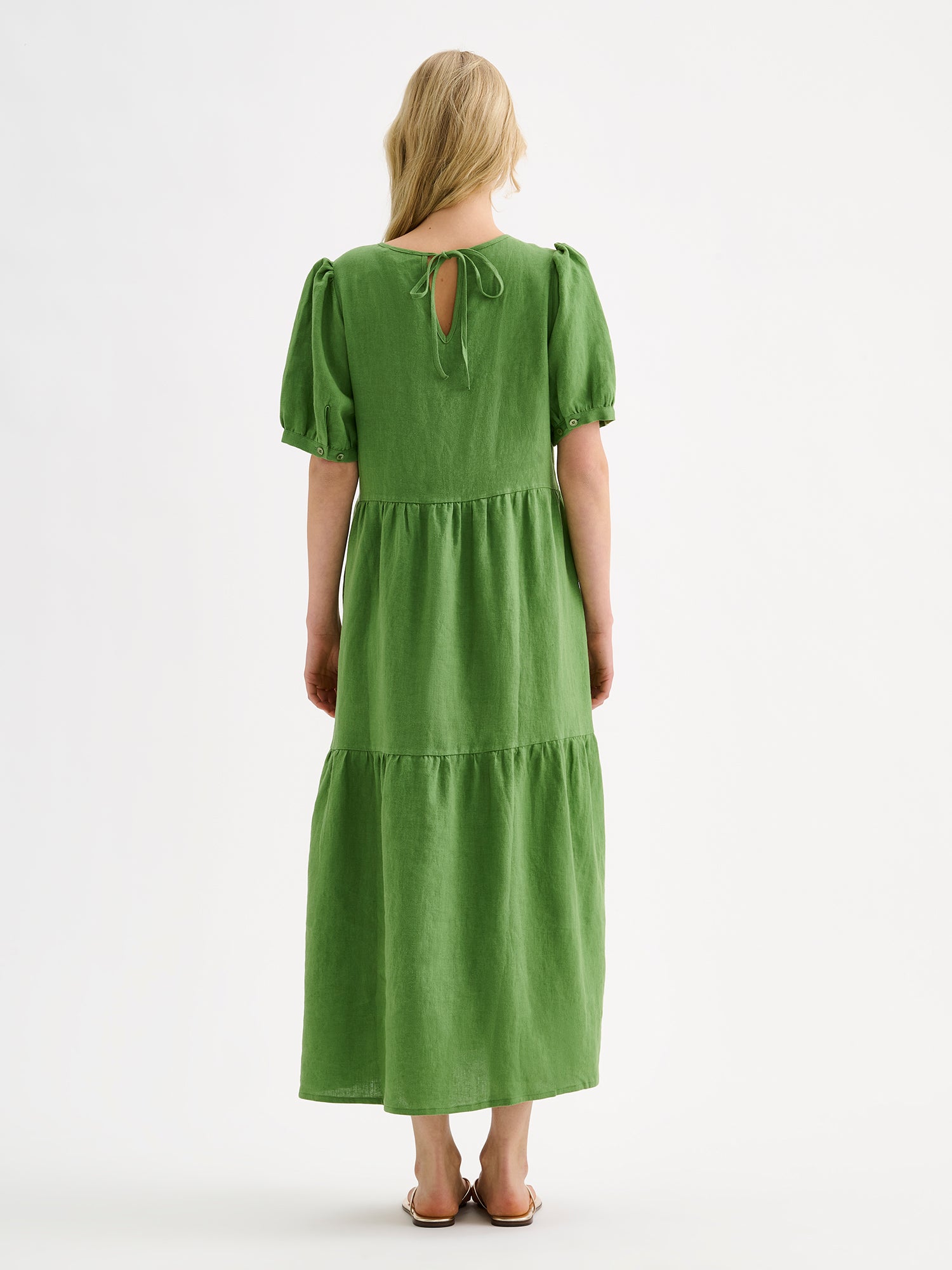 The Sarastus green dress for women is made of high-quality, durable, and breathable woven linen fabric. With its voluminous frill layers and short puff sleeves, this dress exudes both elegance and comfort. Pictured on a blonde model, from the backside