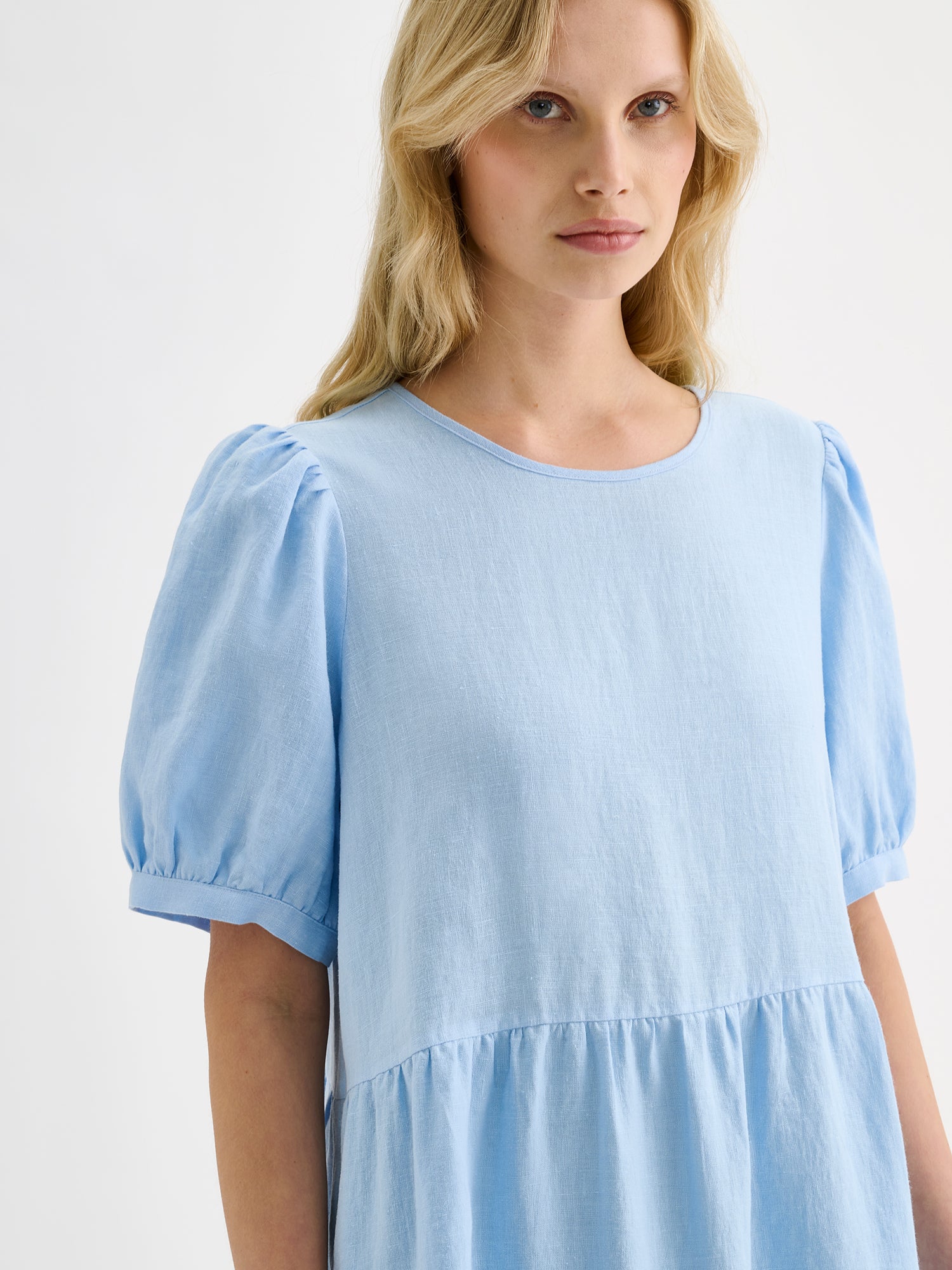The Sarastus long soft light blue dress for women is made of high-quality, durable, and breathable woven linen fabric. With its voluminous frill layers and short puff sleeves, this dress exudes both elegance and comfort. Pictured on a blonde model, close-up.