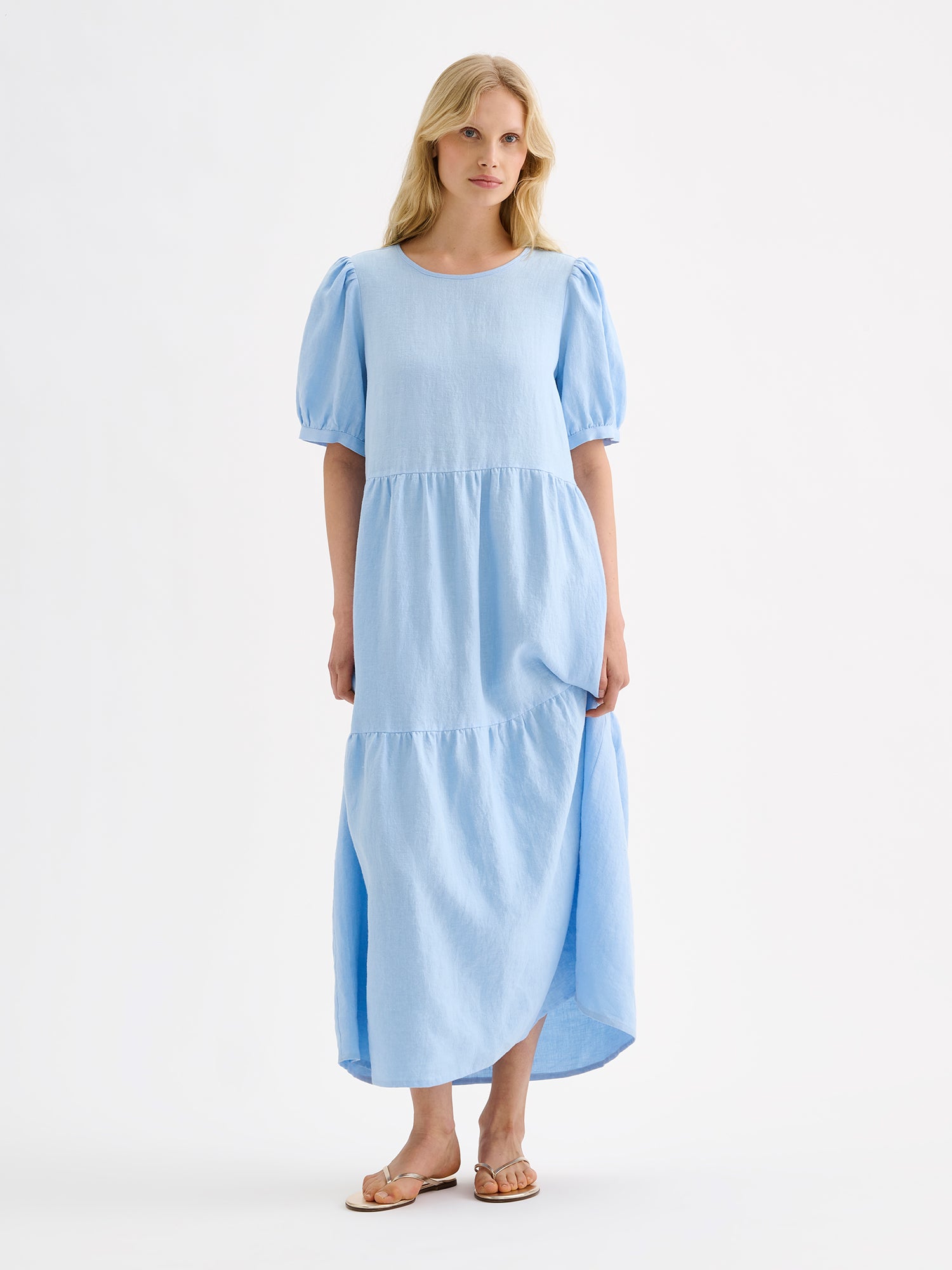 The Sarastus long soft light blue dress for women is made of high-quality, durable, and breathable woven linen fabric. With its voluminous frill layers and short puff sleeves, this dress exudes both elegance and comfort. Pictured on a blonde model.