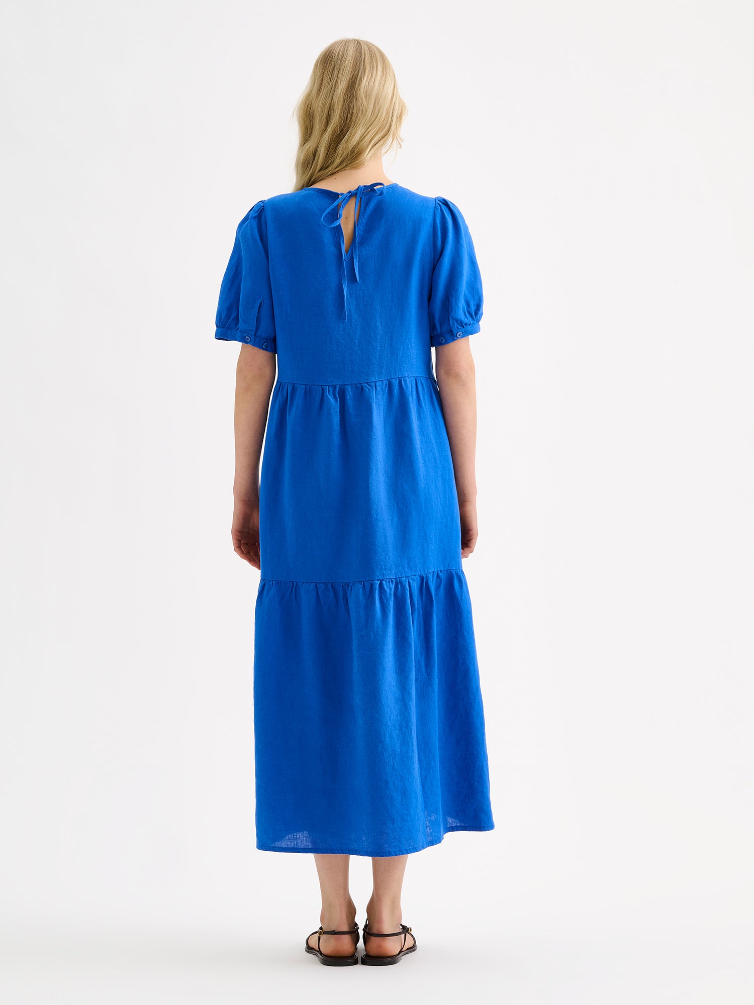 The Sarastus long blue dress for women is made of high-quality, durable, and breathable woven linen fabric, on a model with sandals, pictured from the backside.