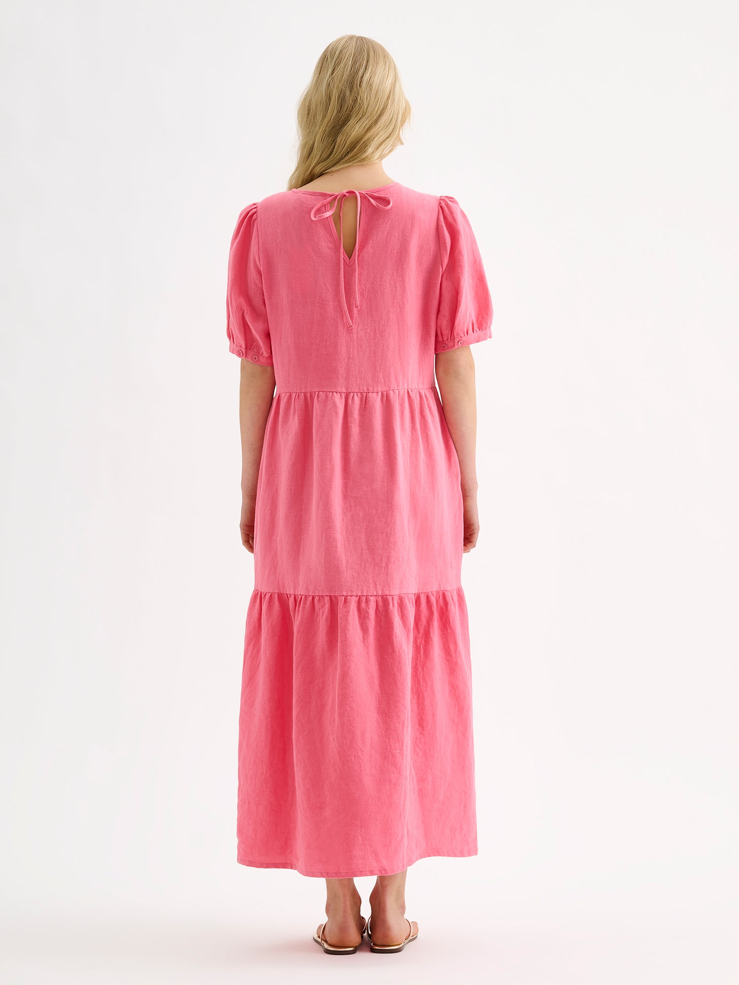 The Sarastus pink dress for women is made of high-quality, durable, and breathable woven linen fabric. With its voluminous frill layers and short puff sleeves, this dress exudes both elegance and comfort. Pictured on a blonde model from the backside