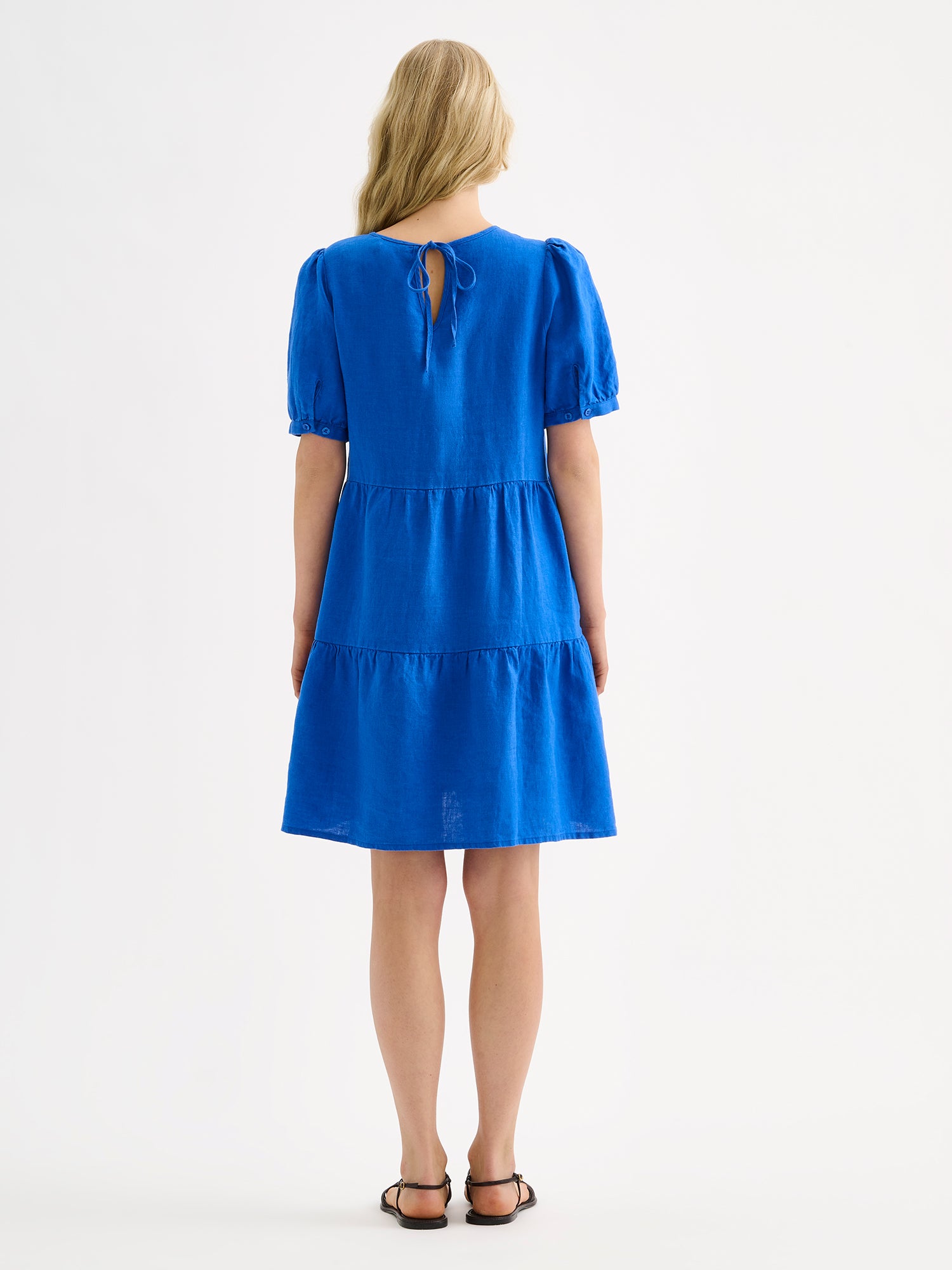 Nanso women's beautiful blue Sarastus linen dress with voluminous frill layers and short puff sleeves, on a model with sandals, pictured from the backside.