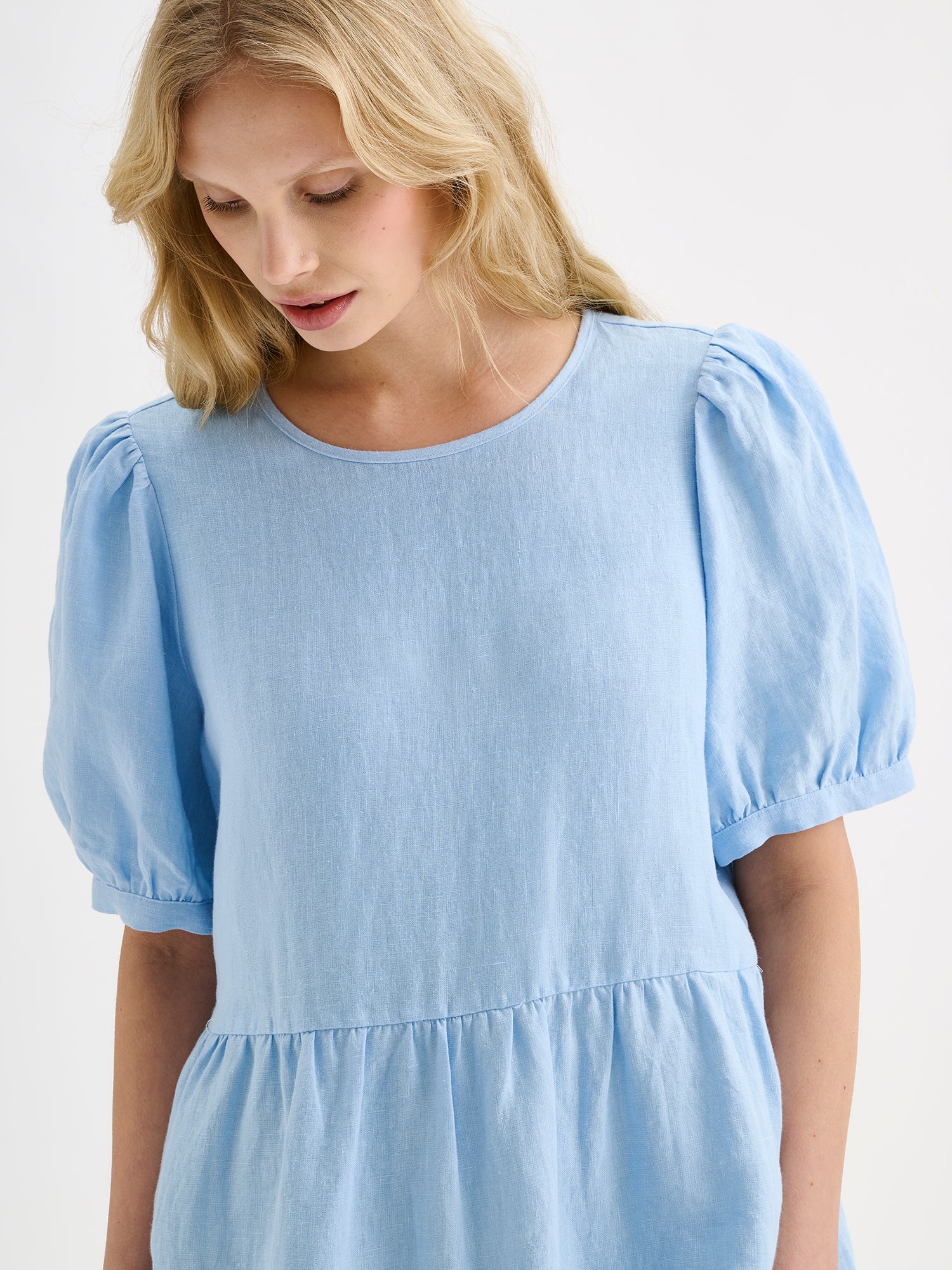 Nanso women's beautiful soft light blue Sarastus midi linen dress features voluminous frill layers and short puff sleeves. This stylish summer dress is made of high-quality linen. Pictured on a blonde model, close-up.
