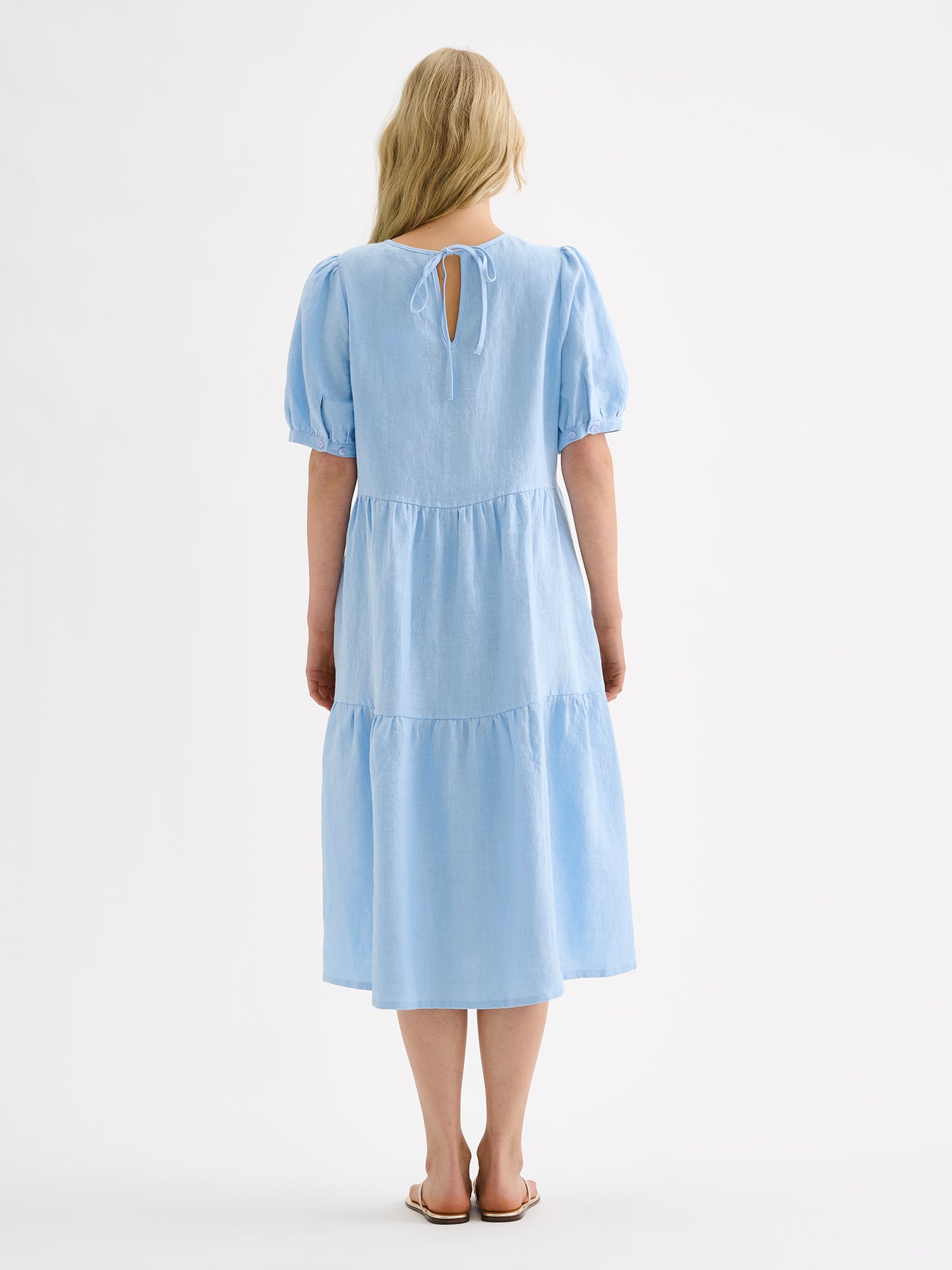 Nanso women's beautiful soft light blue Sarastus midi linen dress features voluminous frill layers and short puff sleeves. This stylish summer dress is made of high-quality linen. Pictured on a blonde model with sandals, from the backside