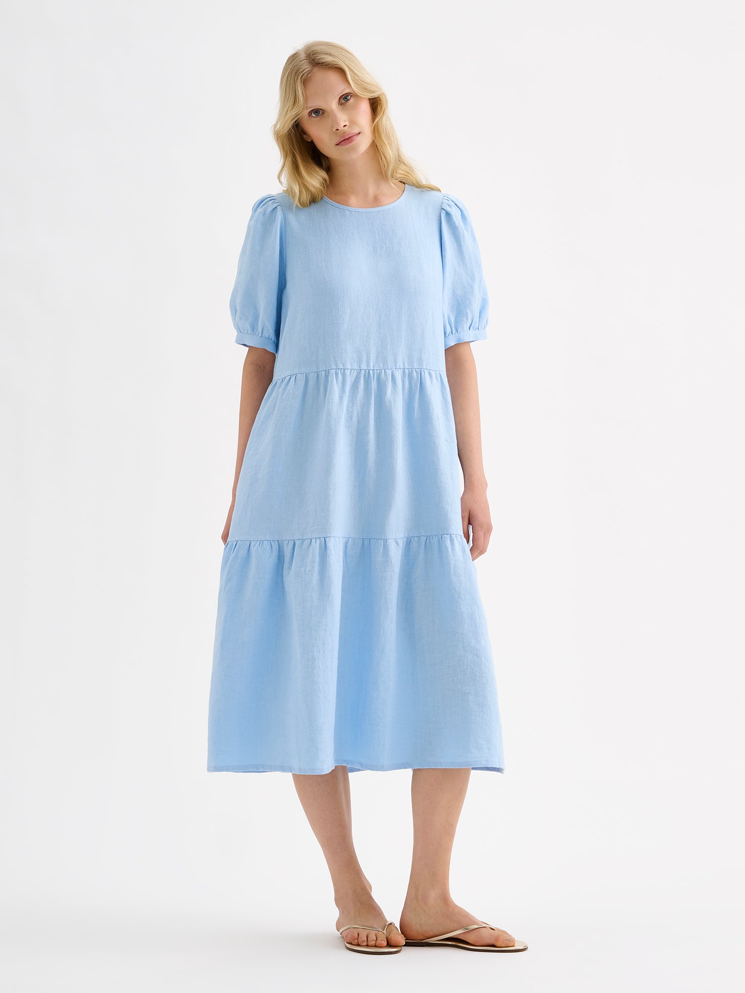 Nanso women's beautiful soft light blue Sarastus midi linen dress features voluminous frill layers and short puff sleeves. This stylish summer dress is made of high-quality linen. Pictured on a blonde model with sandals.
