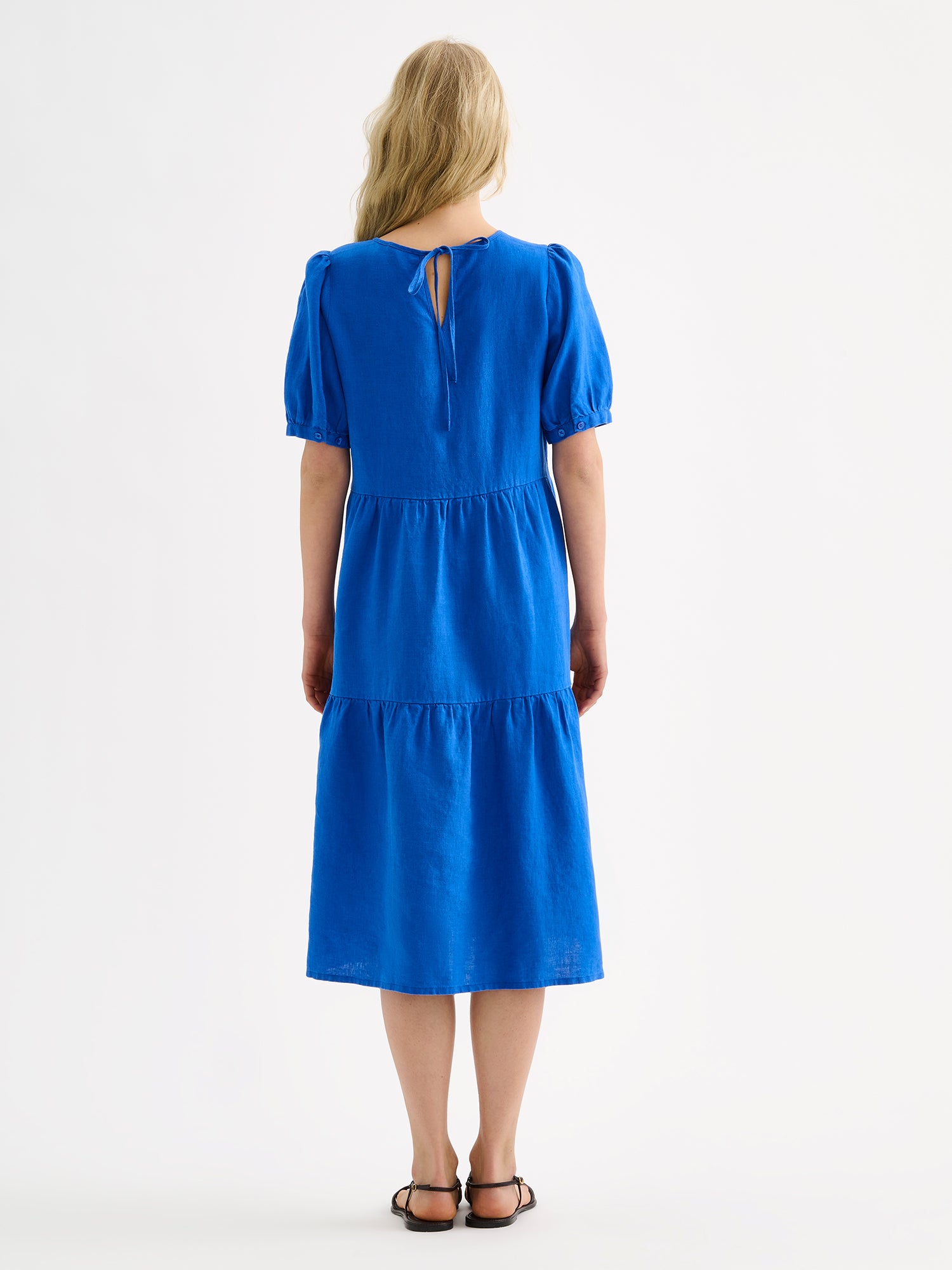Nanso women's beautiful blue Sarastus midi linen dress features voluminous frill layers and short puff sleeves, on a model with sandals, pictured from the backside.