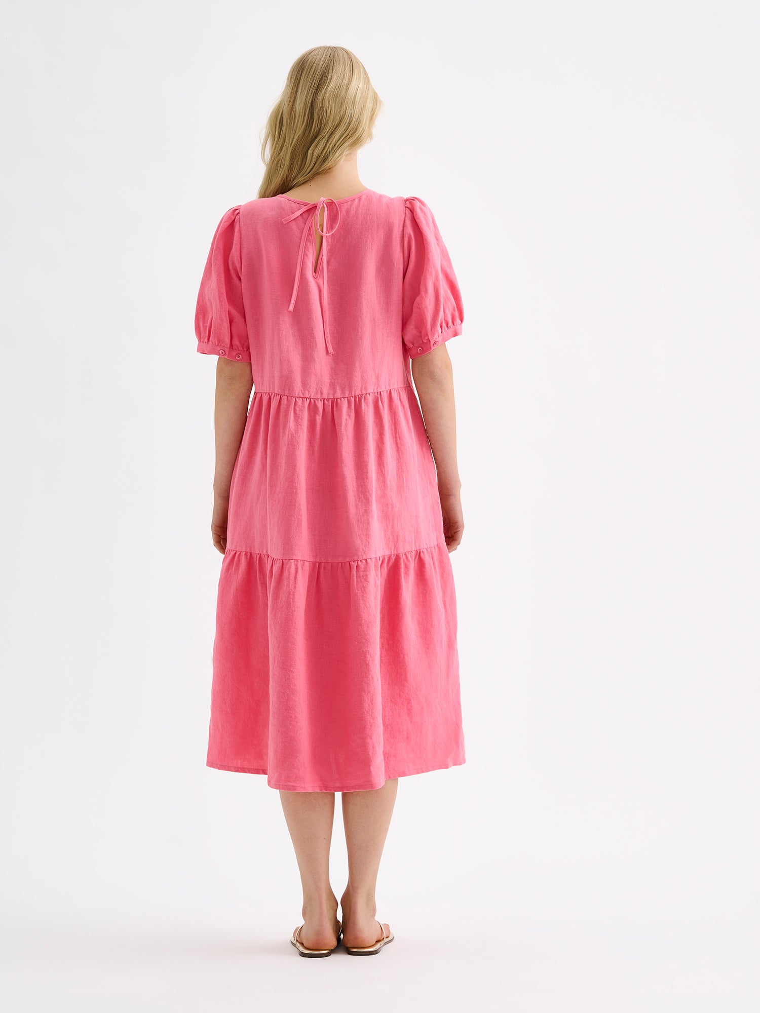 Nanso women's beautiful pink Sarastus midi linen dress features voluminous frill layers and short puff sleeves. This stylish summer dress is made of high-quality linen. Pictured on a blonde model with sandals, from the backside
