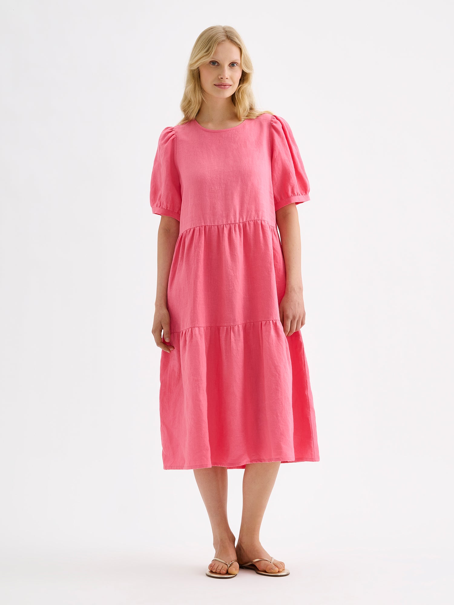 Nanso women's beautiful pink Sarastus midi linen dress features voluminous frill layers and short puff sleeves. This stylish summer dress is made of high-quality linen. Pictured on a blonde model with sandals.