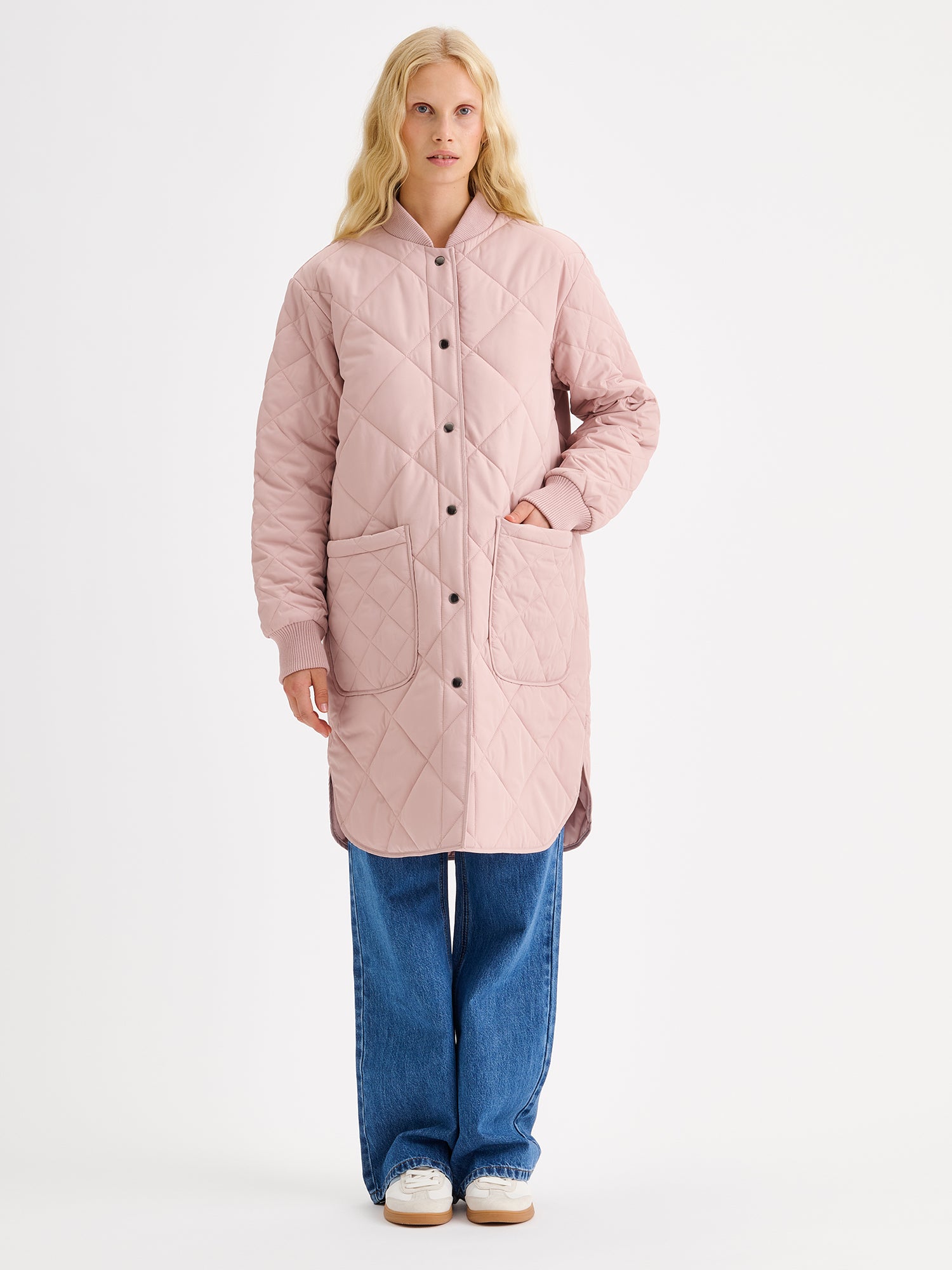 Nanso women's casual Midi quilted coat in dark blue features lightweight padding, ribbed knit detailing at the neckline and cuffs, snap closures and front pockets. On a model with jeans and sneakers.