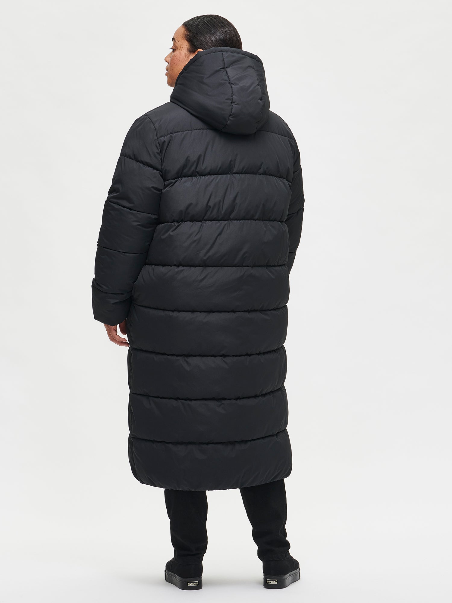 The Lumi Long Pink Puffer Coat in black offers cozy warmth with its lightweight, puffy insulation. Worn by a model from the back.