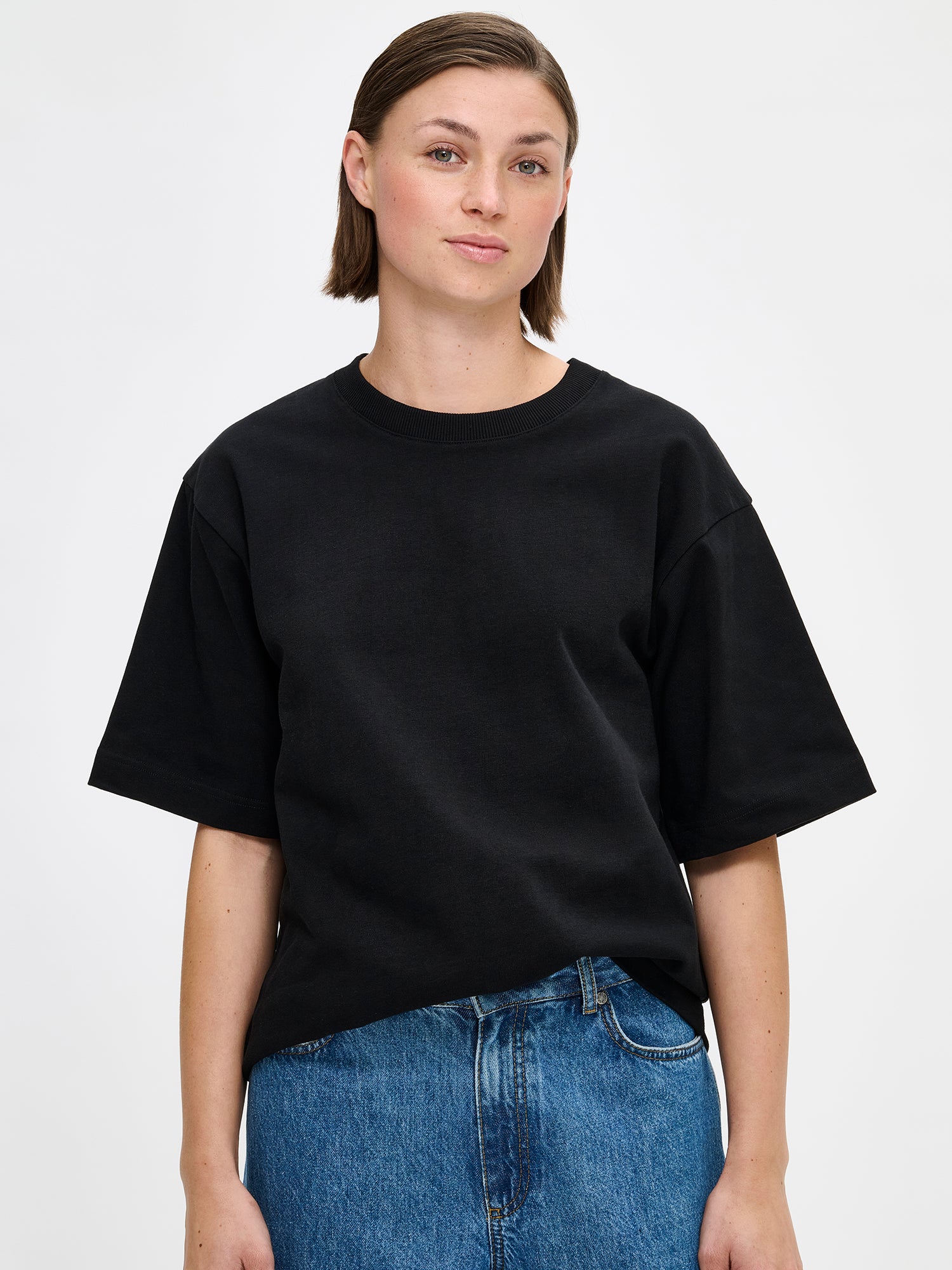 Nanso women's solid black Forma T-shirt. This boxy, loose-fitting T-shirt features a round ribbed neckline and loose 2/4-length sleeves. On a model with relaxed jeans, close-up.