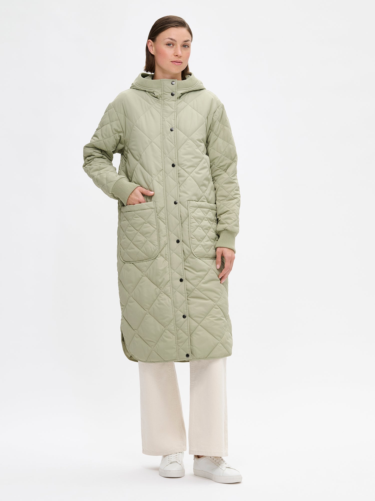 Nanso women's casual green Maxi quilted coat with light padding and a cozy hood. Perfect for in-between seasons, the hooded jacket features a two-way front zipper with a snap-button wind flap, a comfortable ribbed hood edge and cuffs. On a model with jeans and sneakers.