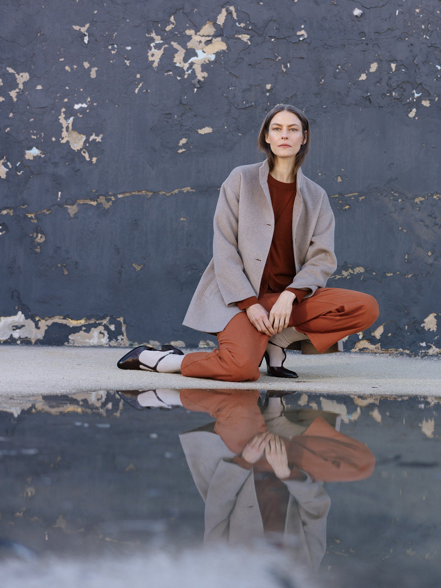 Woman in a beige coat and red pants sitting on a reflective surface with a textured wall background