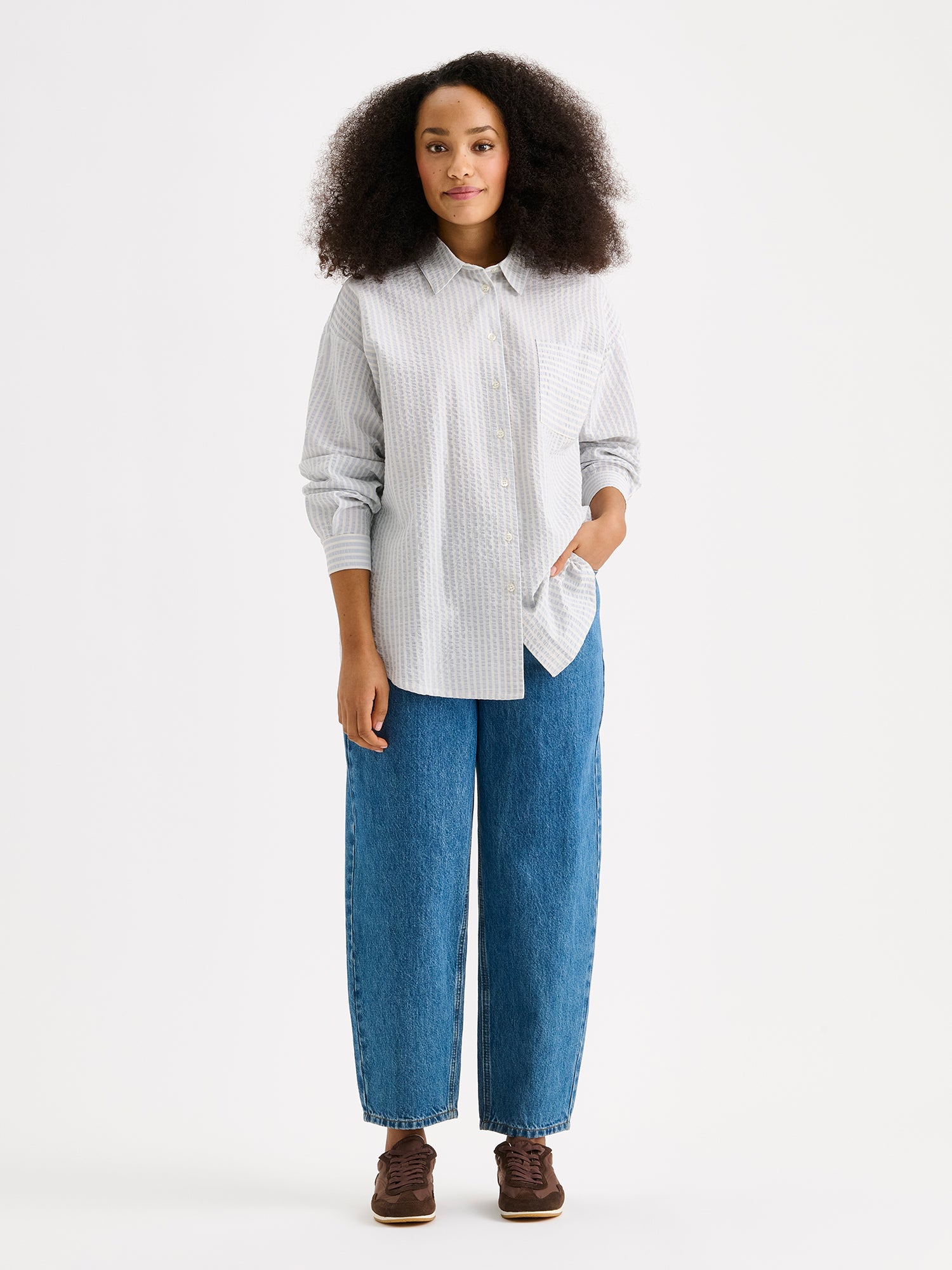 Nanso women's Marea Shirt in pale blue features a classic button-up shirt with a timeless and striped pattern. The shirt includes a collar, dropped shoulder line, chest pocket, and front buttons. On a model with jeans.
