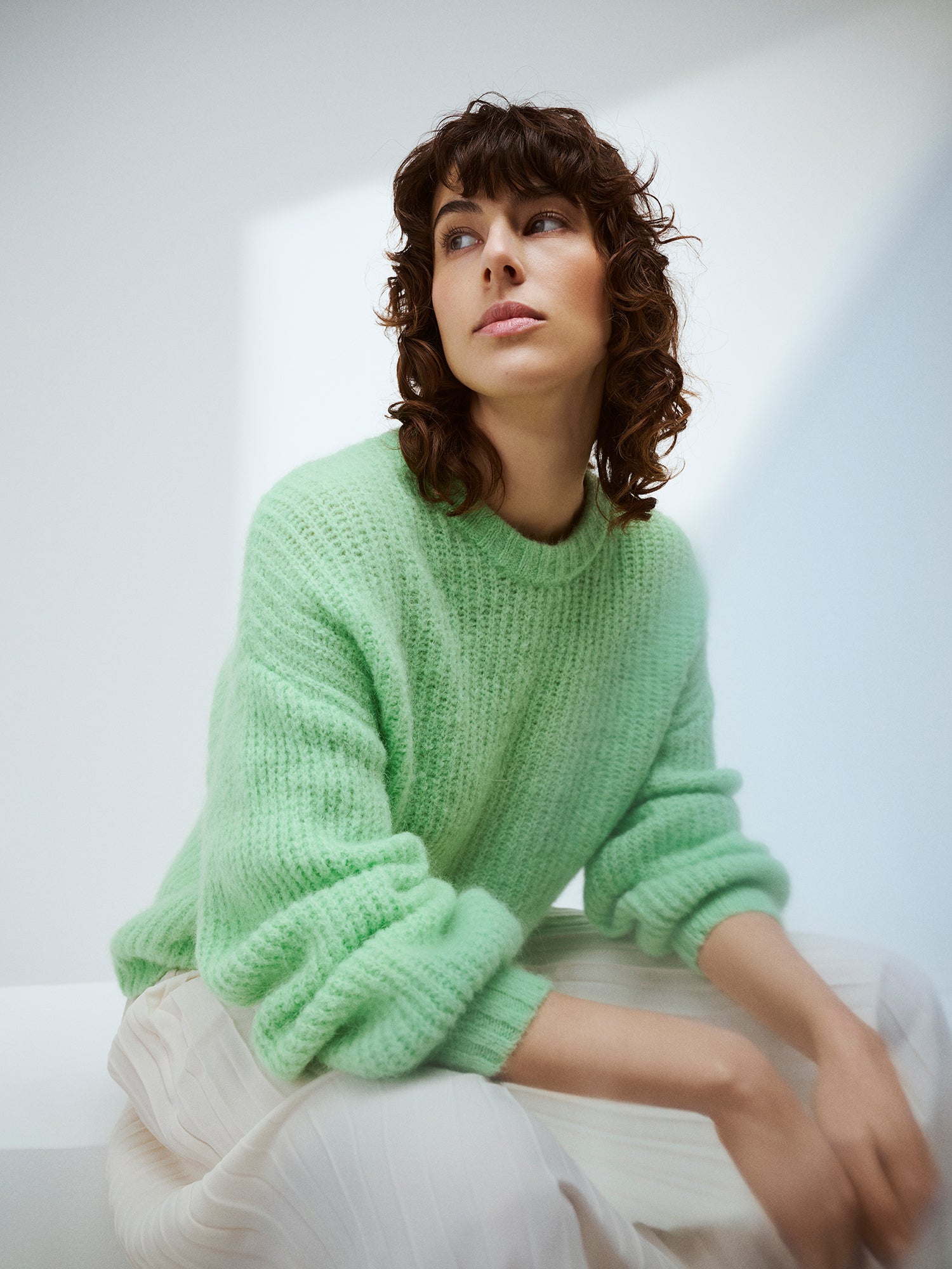 KARAMELLI women's luxurious mohair blend Knit Sweater in light green shade features a round neckline and full-length sleeves. Pictured on a sitting model with a white skirt.