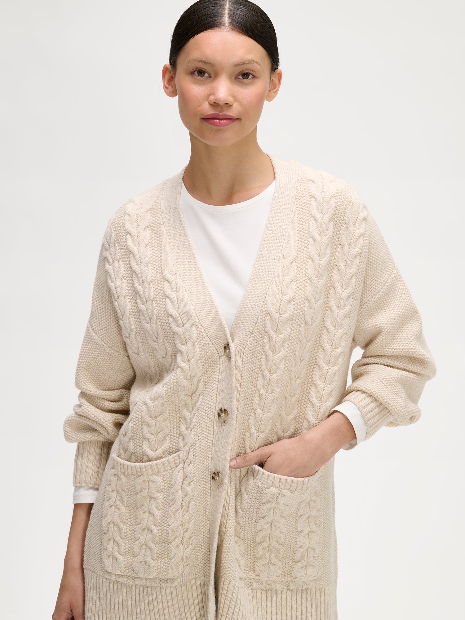 Woman wearing a beige cable knit cardigan over a white shirt on a light gray background