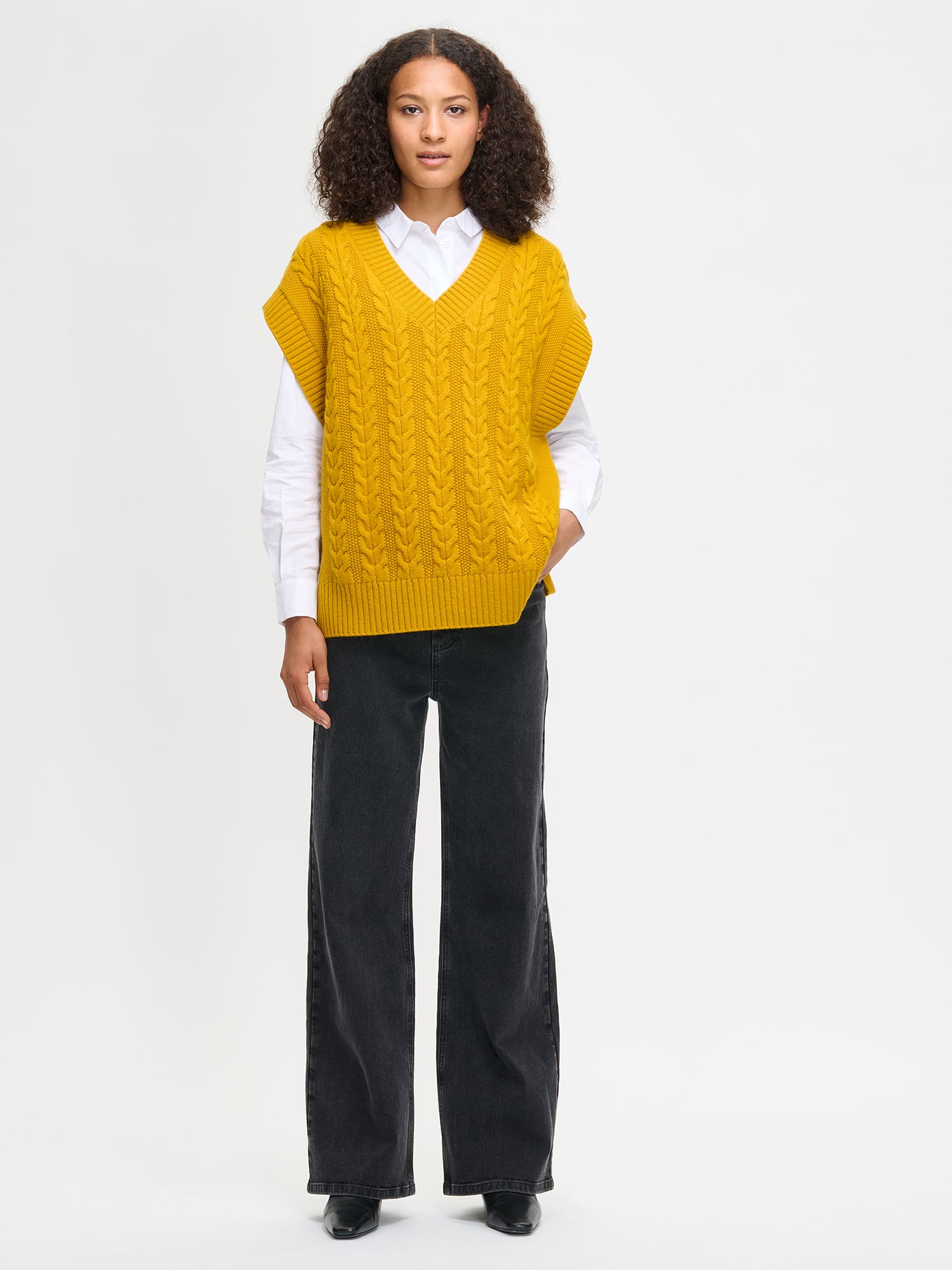 Woman wearing a yellow knitted vest over a white shirt with black pants on a light gray background