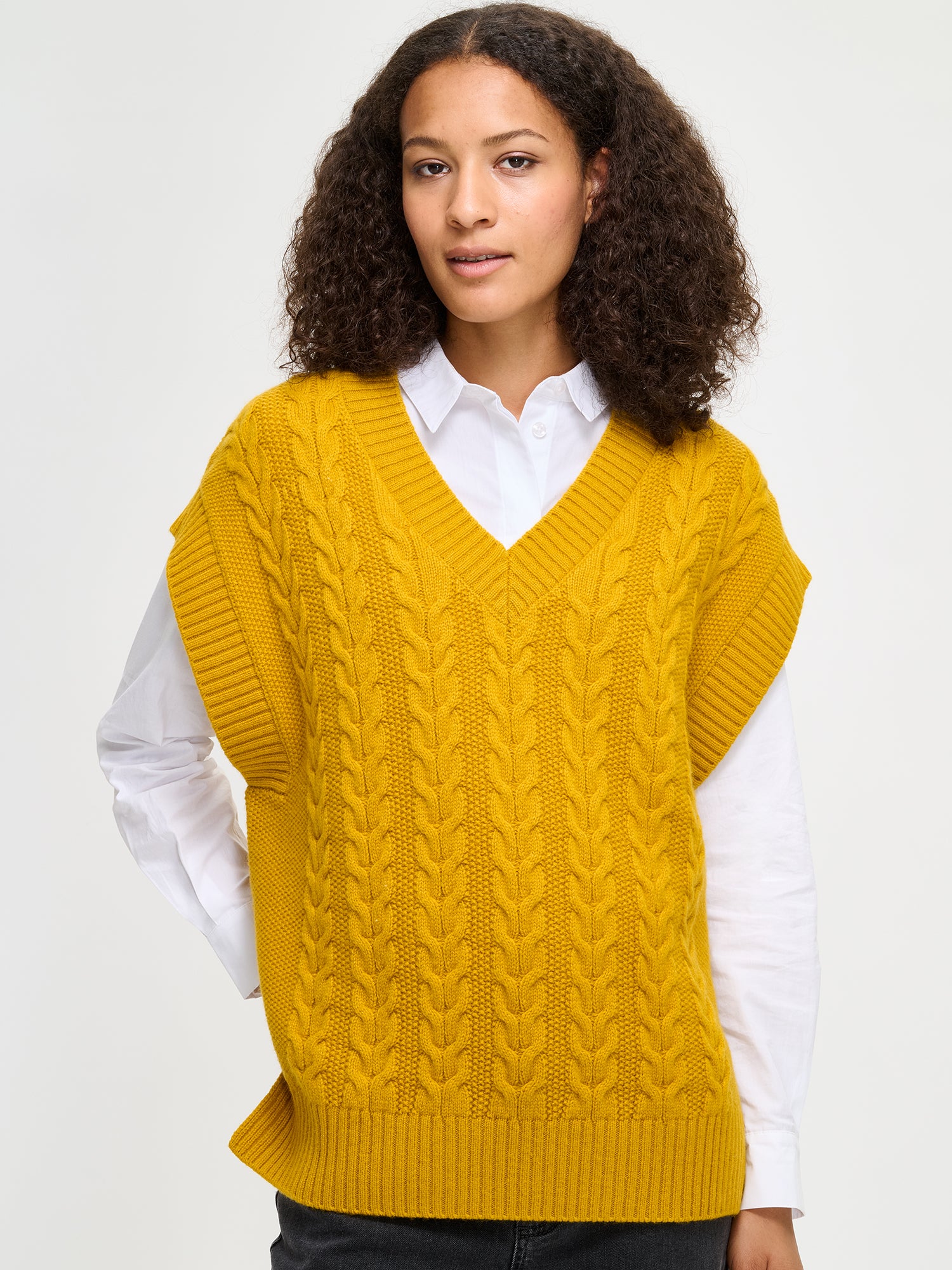 Woman wearing a yellow cable knit sweater vest over a white shirt on a light gray background