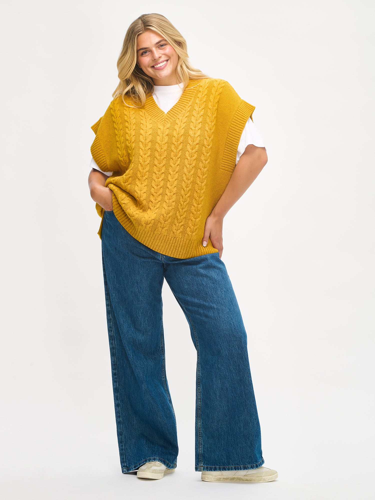 Woman wearing a yellow knit vest and blue jeans on a white background