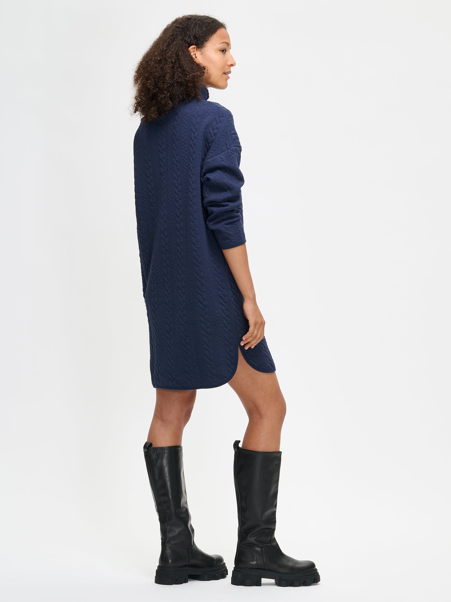 Woman wearing a dark blue dress with a turtleneck and black boots on a white background