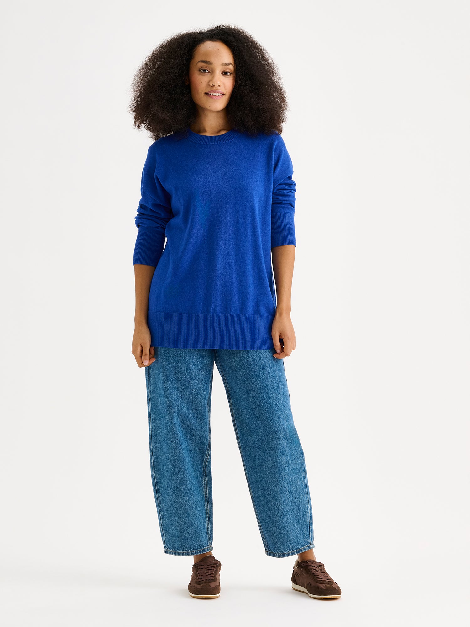 The VILLIS Long Knit Sweater in blue features a timeless boxy silhouette and a classic crew neckline, dropped shoulders, and full-length sleeves and ribbed trim at the hem and cuffs. On a model with blue jeans.