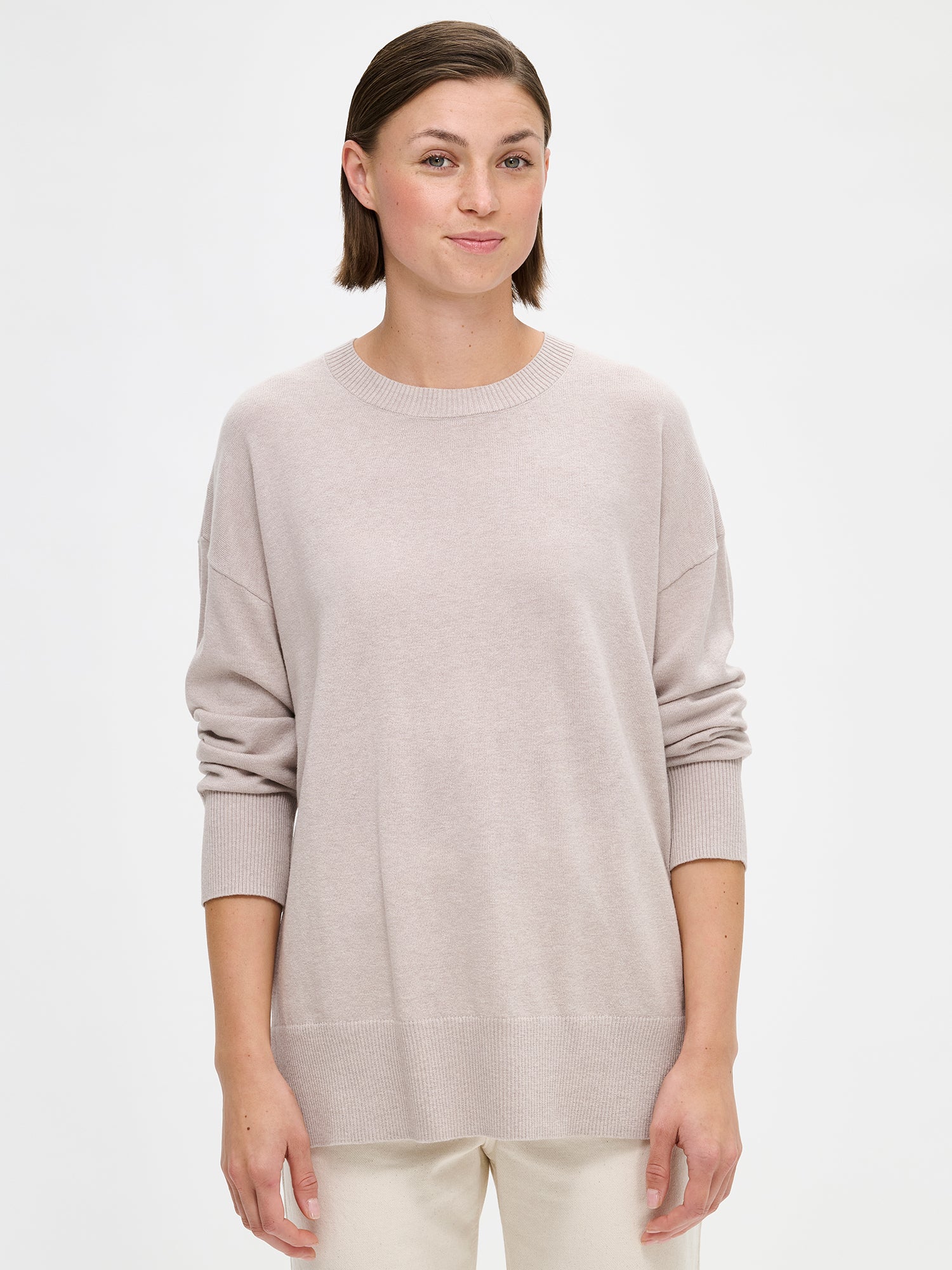 The VILLIS Long Knit Sweater in greige features a timeless boxy silhouette and a classic crew neckline, dropped shoulders, and full-length sleeves and ribbed trim at the hem and cuffs. On a model with off white jeans and sneakers, close-up.