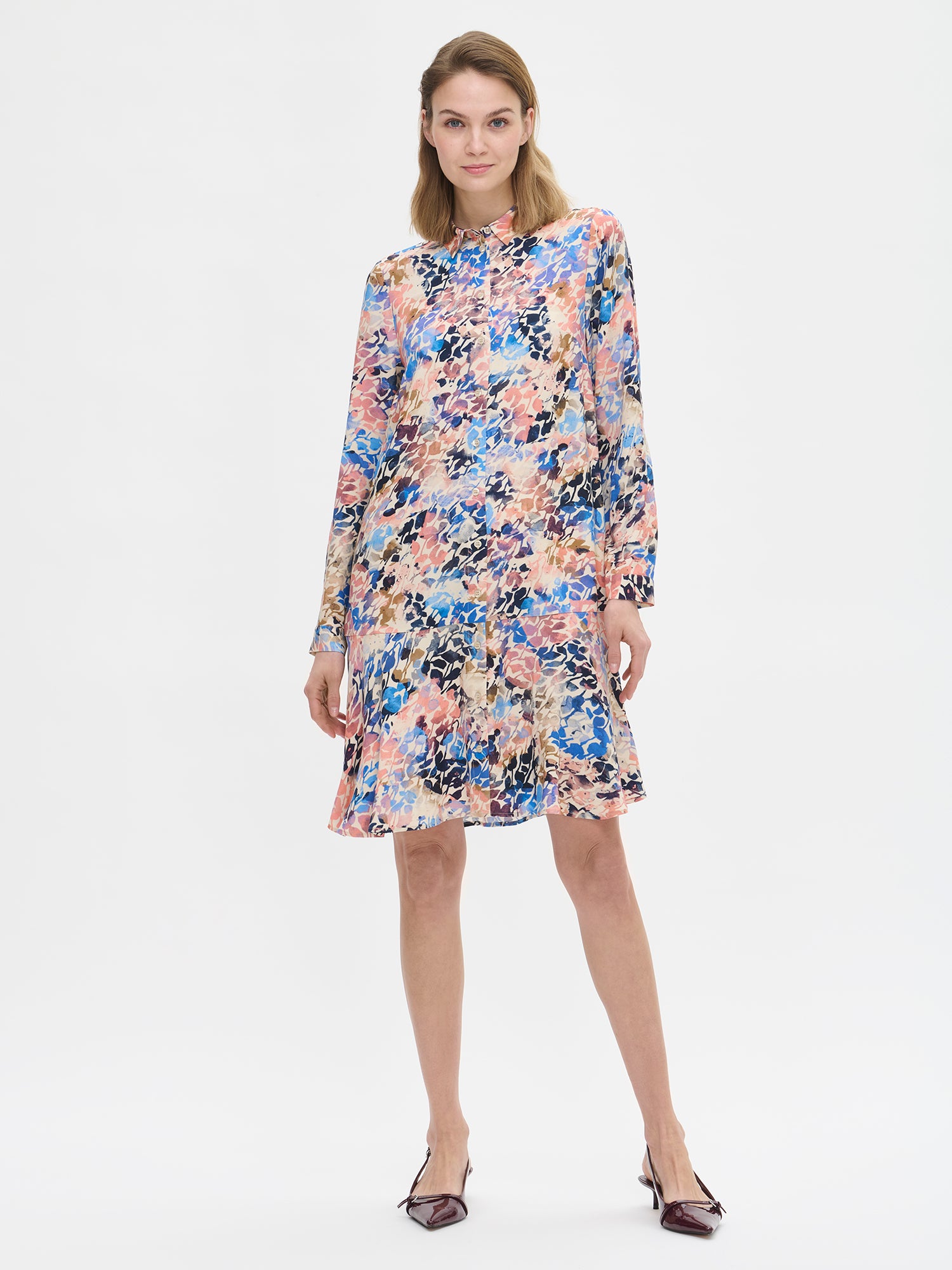 Women's SAGITTA Dress with vibrant pattern and flared hem. The dress features a front button closure and timeless collar design, paired with full-length sleeves. Pictured on a model with high heels.