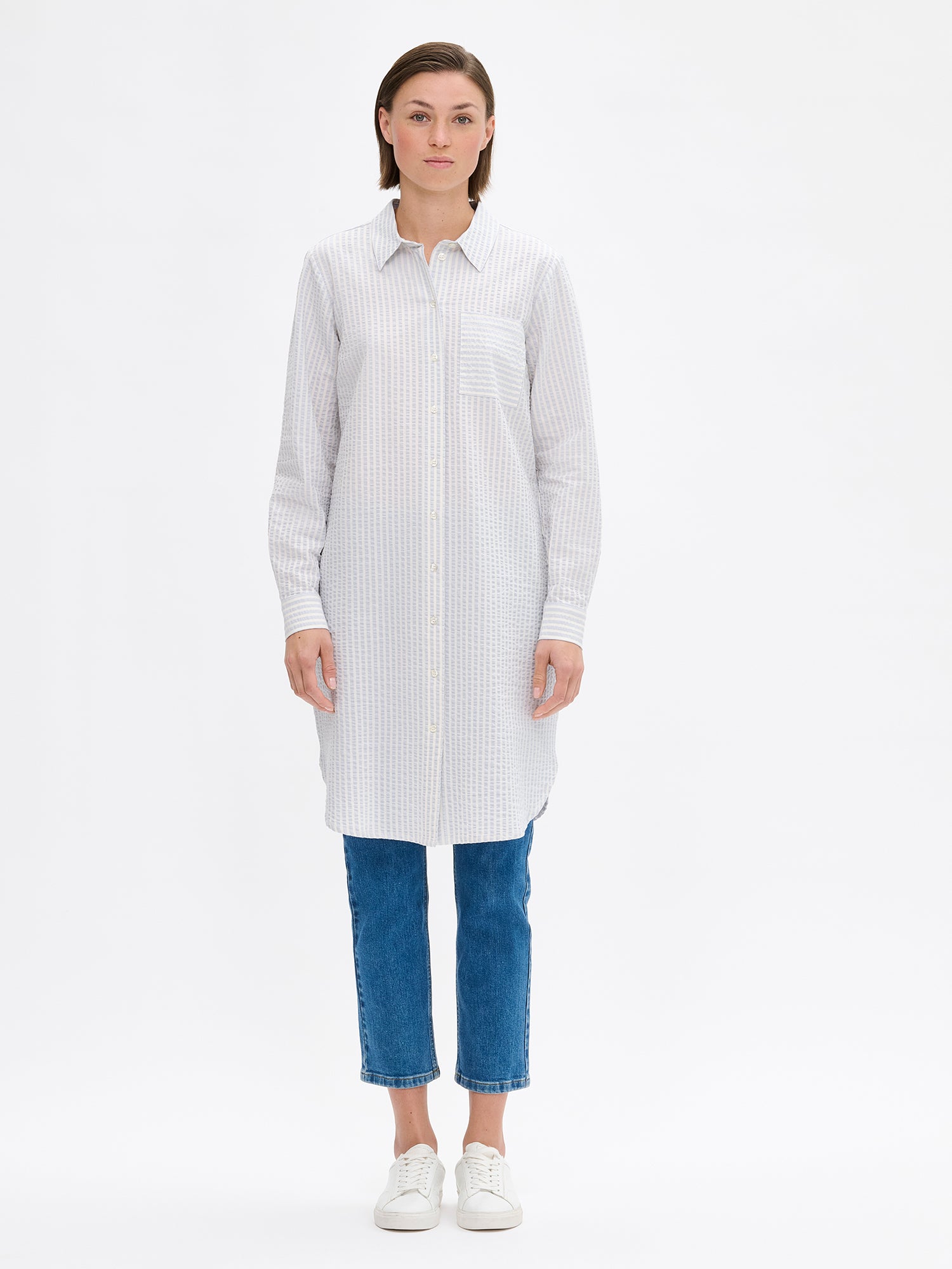 Nanso women's MAREA shirt dress in pale blue features a classic collar, front button closure, rounded hemline. A chest pocket and side pockets add everyday functionality. On a model with jeans and sneakers.
