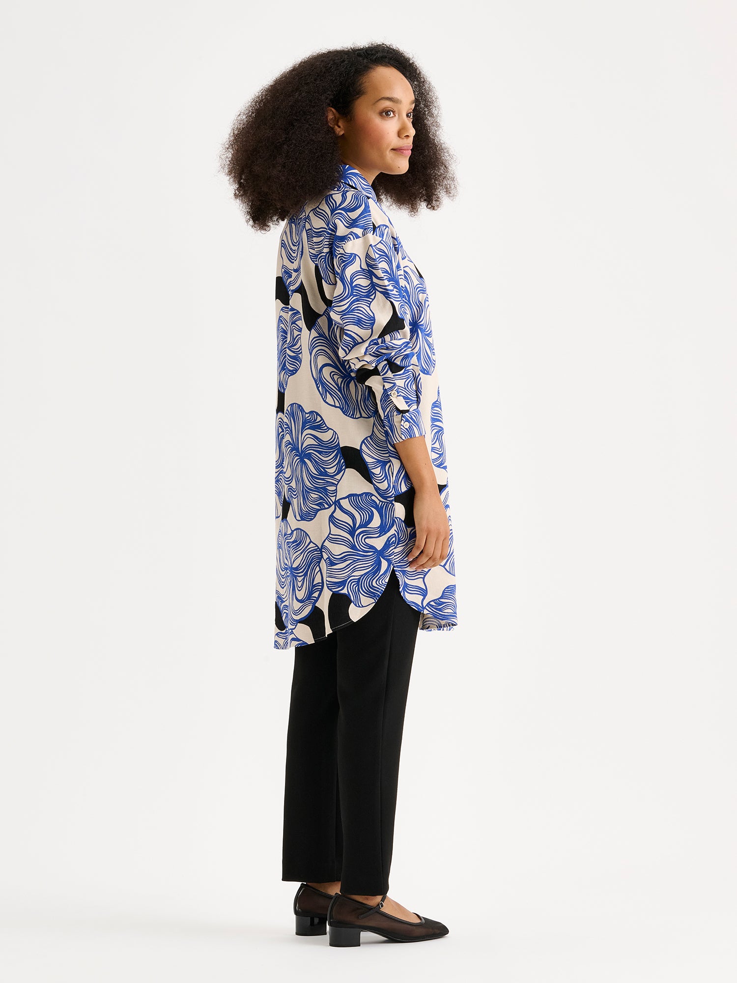Women's KOSMO shirt features a striking blue patterned design paired with a timeless collar style, full-length sleeves, a practical front button closure and side pockets. On a model with black trousers photographed from the side.