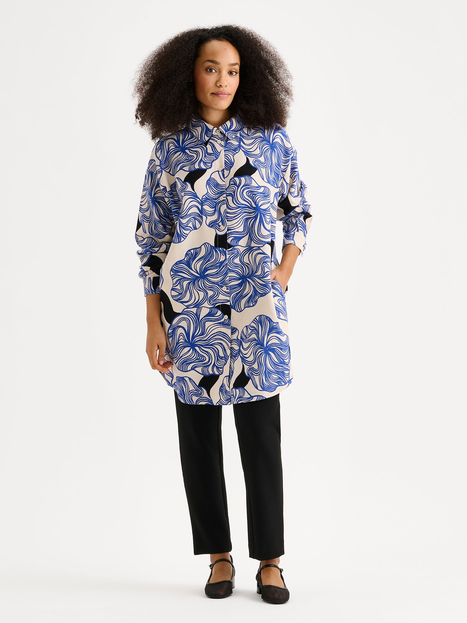 Women's KOSMO shirt features a striking blue patterned design paired with a timeless collar style, full-length sleeves, a practical front button closure and side pockets. On a model with black trousers.