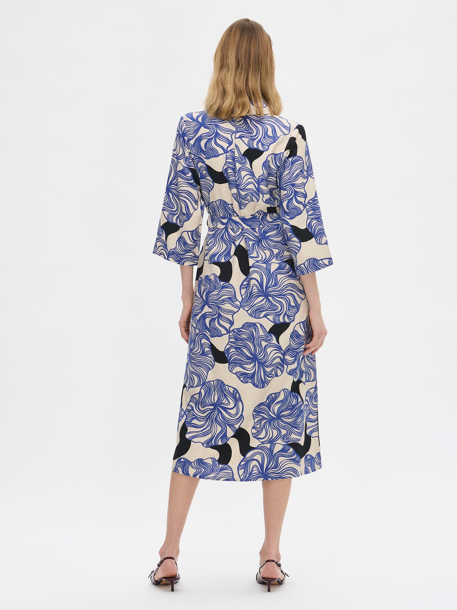 Nanso KOSMO Dress combines timeless elegance with a striking blue pattern, adjustable waist belt for a customized fit and practical pockets. On a model with high heels photographed from the back side.