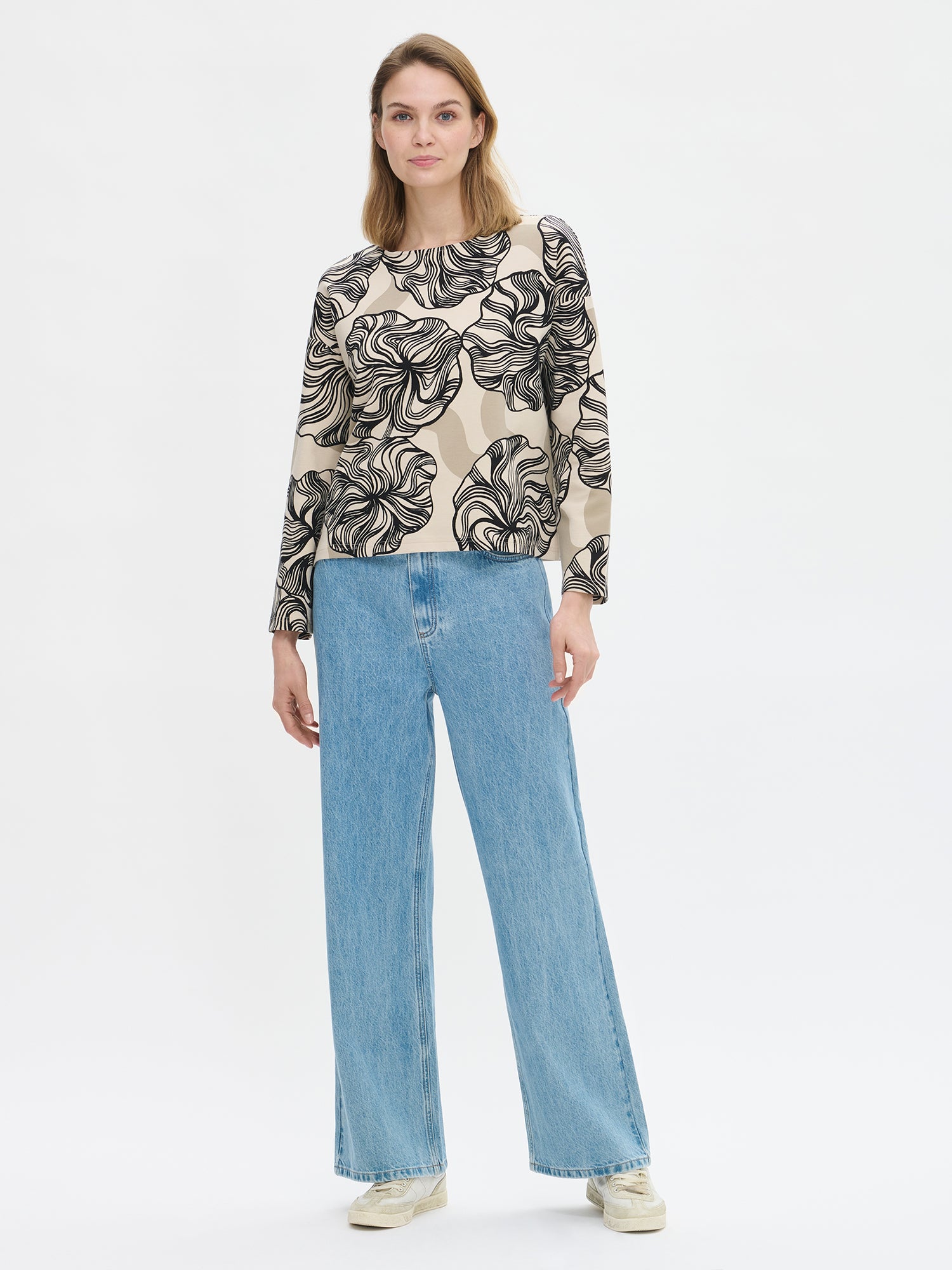 The Kosmo patterned blouse for women with a boat neckline, dropped shoulders, and full-length sleeves and side slits at the hem. On a model with jeans.
