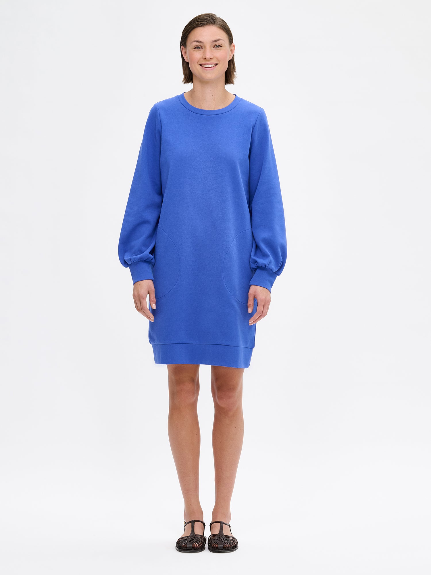 Nanso women's blue Leija-dress. This straight-cut dress features a round neckline, side pockets, and full-length sleeves that gather beautifully at the cuffs. On a model with sandals.