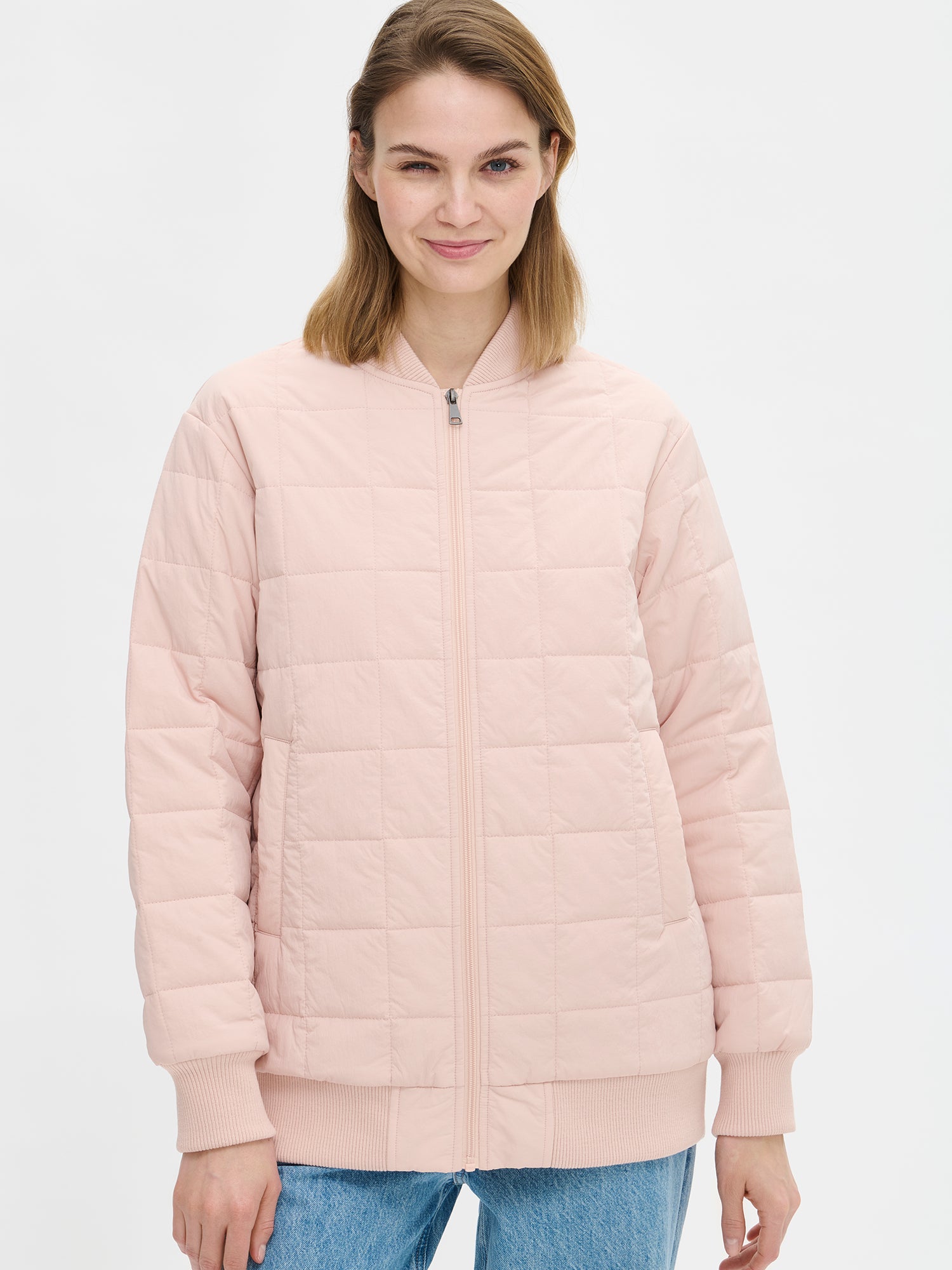 Nanso MERJE Quilted Bomber Jacket in Light Rose features a lightweight Sorona -insulation, ribbed knit detailing at the neckline, cuffs, and hem. Dropped shoulders create a relaxed, contemporary silhouette. Photographed close-up on a model with jeans on, jacket closed.