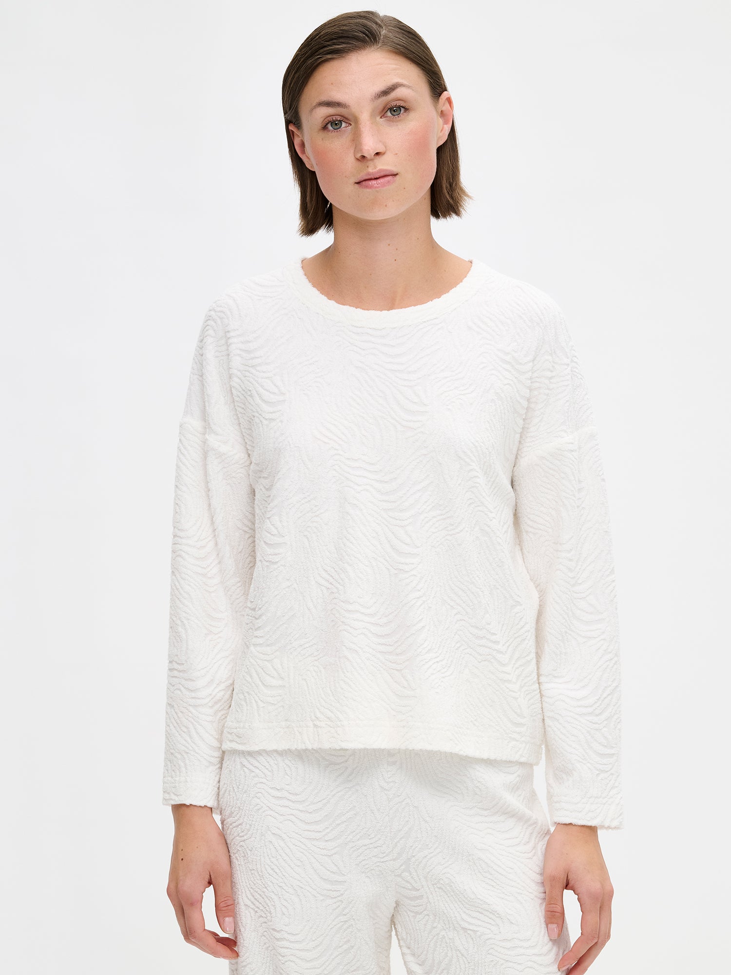 The VÄRE Shirt in off-white features long sleeves and a classic round neckline and a beautifully textured jacquard knit fabric, designed for relaxed comfort. Photographed on a model, close-up.