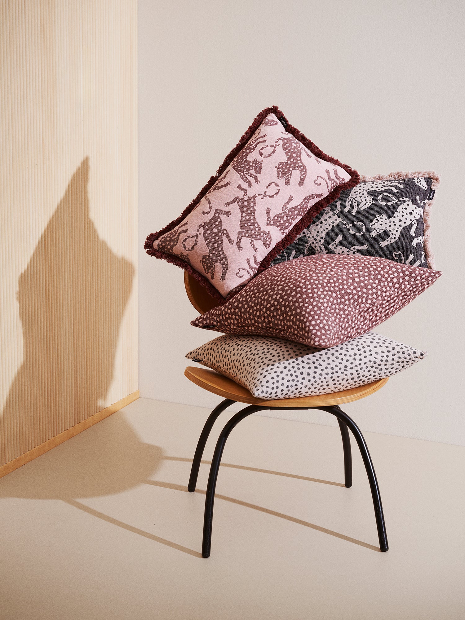 The leopard-print Hapsu pillowcase in a soft rose shade with a black Hapsu pillow case and other same colored pillows on a chair in a beige room.