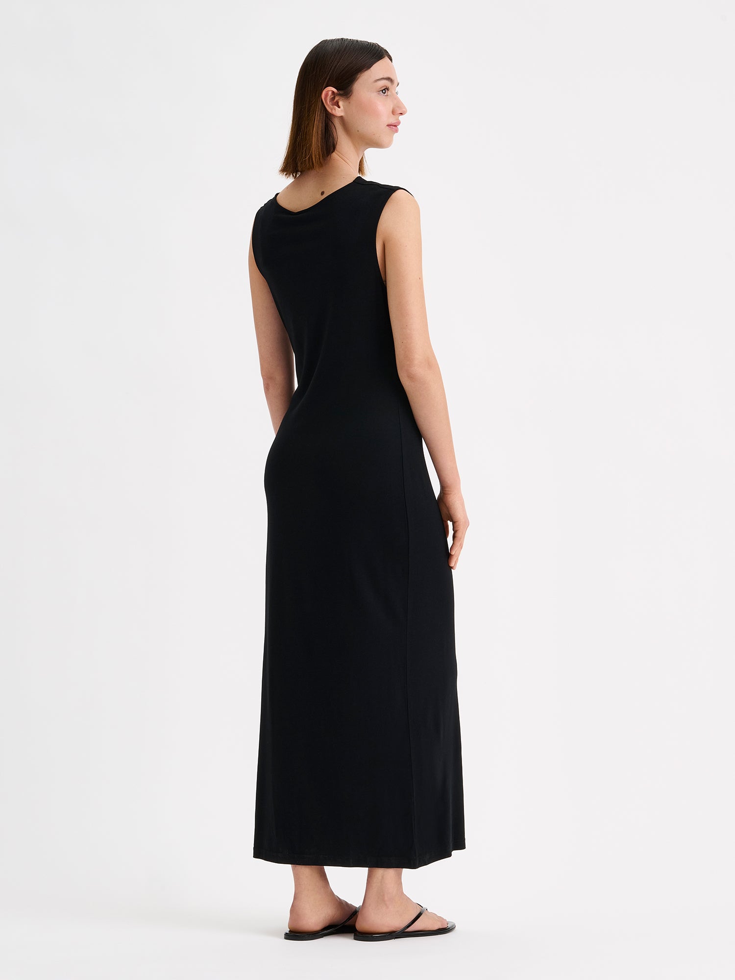 Nanso women's timeless Vivian black dress combines effortless style and comfort. This sleeveless maxi dress features an elegant draped neckline and a subtle side slit. Pictured on a brunette model, from the backside.