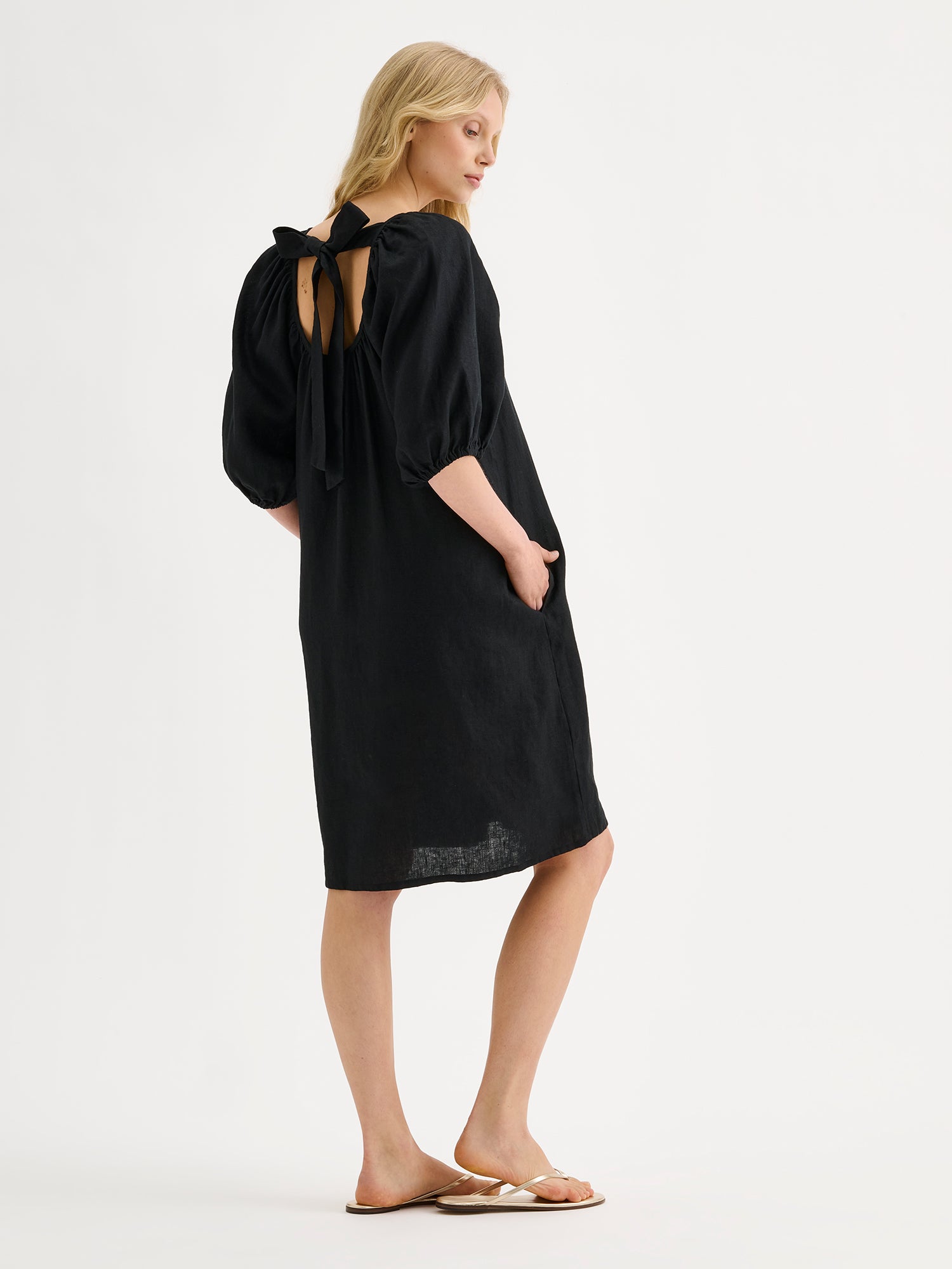 Nanso women’s beautifully relaxed black Donna linen dress. This airy short dress features a softly gathered open back neckline, finished with a tie-back bow detail. Pictured on a model, from the backside with a bow detail showing.