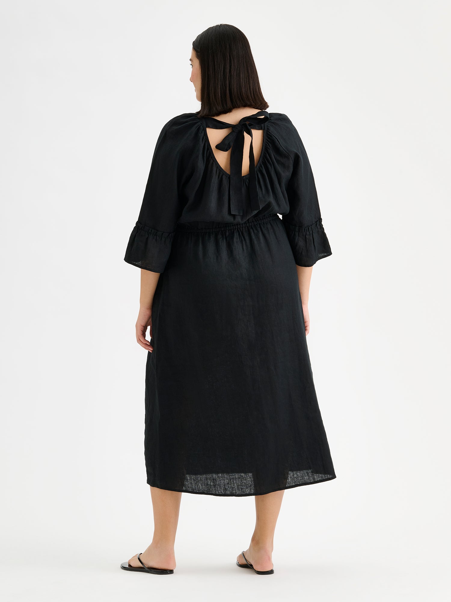 Nanso women's black Donna midi linen dress combines effortless elegance with feminine details. This airy midi-length style features an open, softly gathered back neckline with a striking self-tie bow. Pictured on a brunette model, from the backside