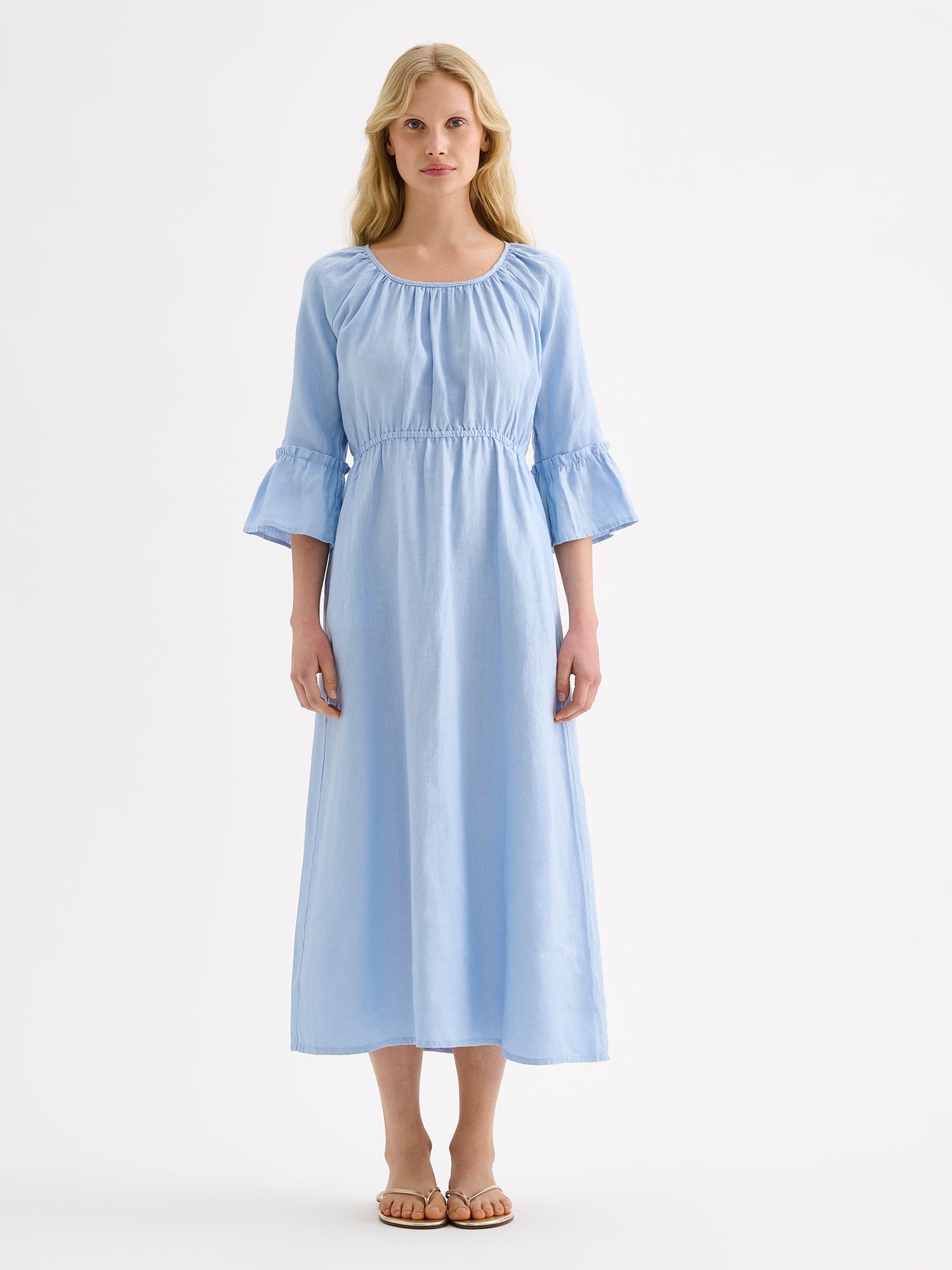 Nanso women's softly light blue Donna midi linen dress combines effortless elegance with feminine details. This airy midi-length style features an open, softly gathered back neckline with a striking self-tie bow. Pictured on a blonde model with black sandals.