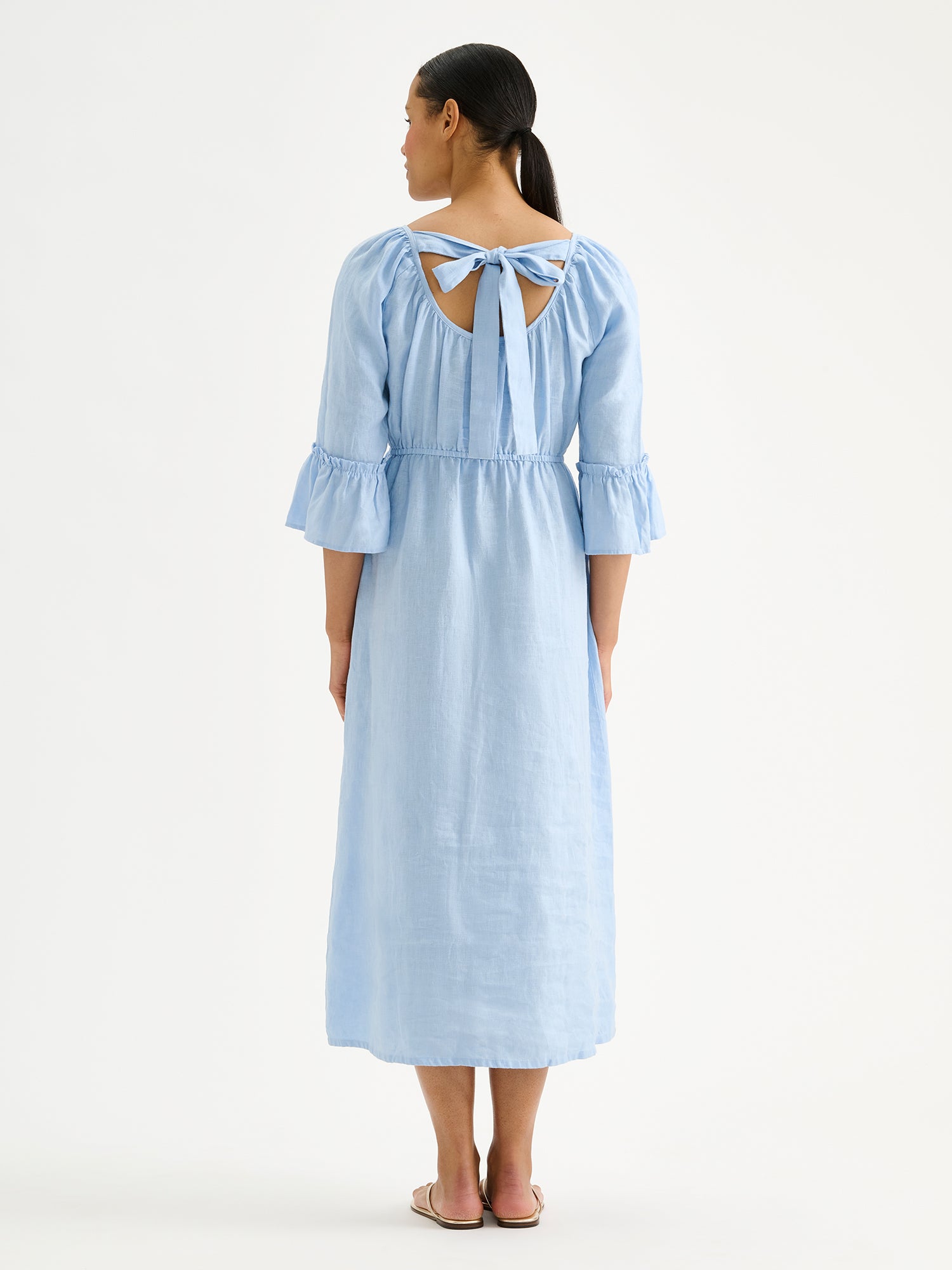 Nanso women's softly light blue Donna midi linen dress combines effortless elegance with feminine details. This airy midi-length style features an open, softly gathered back neckline with a striking self-tie bow. Pictured on a brunette model with golden sandals, from the backside.