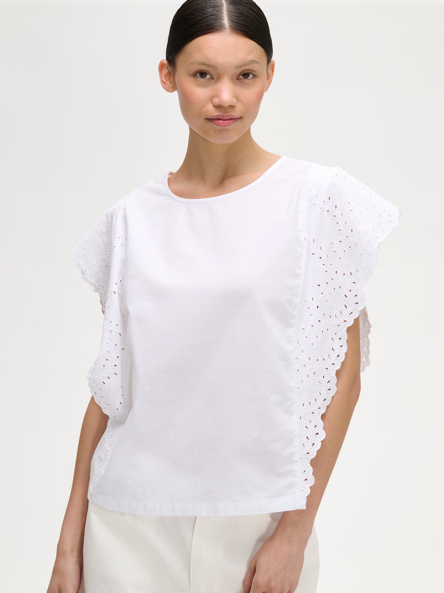 Nanso women's white Sevilla blouse for women combines an airy silhouette with delicate lace details. On a model with white shorts, close-up.