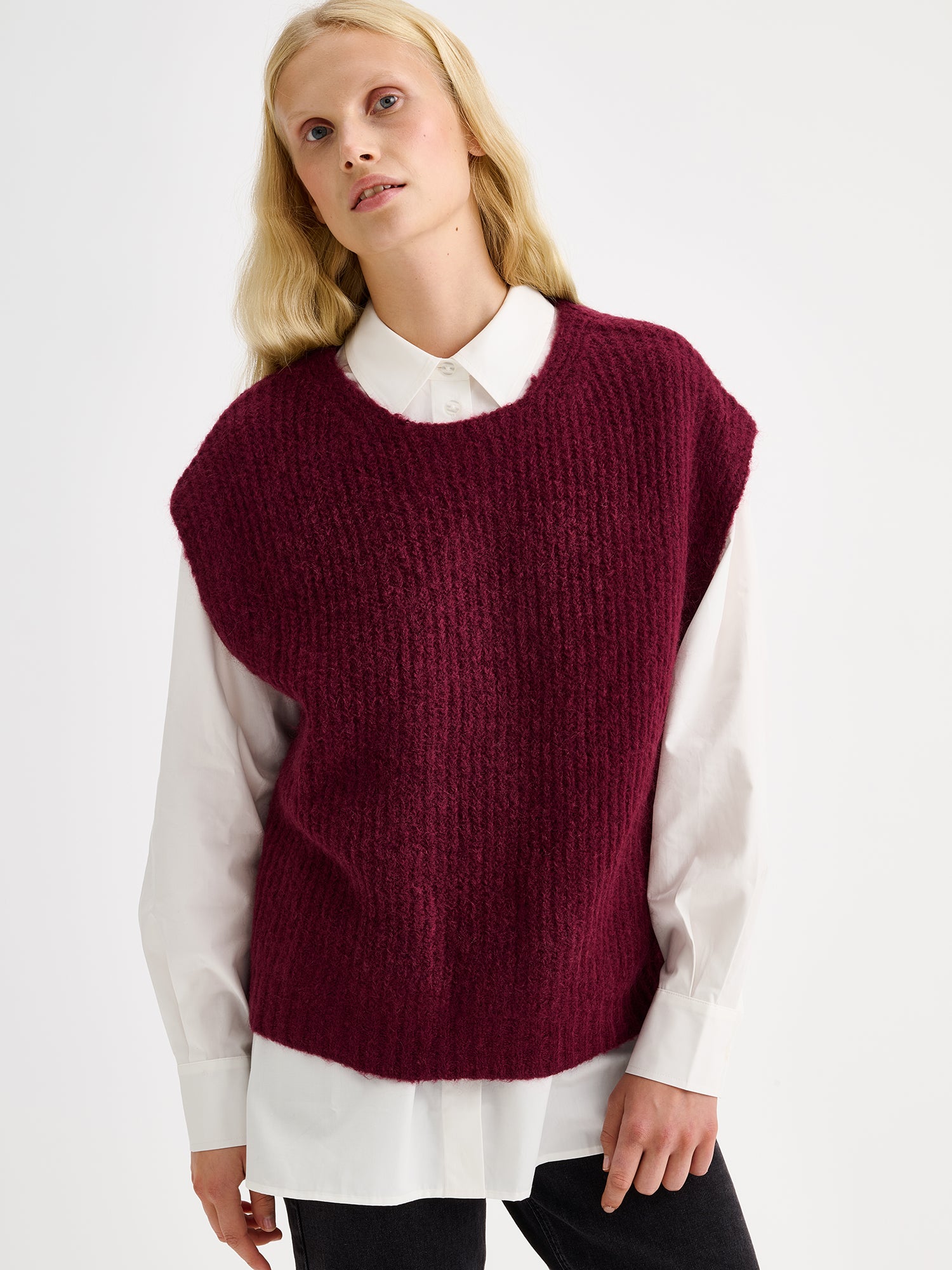 Nanso women's luxuriously soft and wonderfully chunky, the Karamelli knitted vest in a rich wine-red hue on a model with a white shirt and gray jeans, close-up.