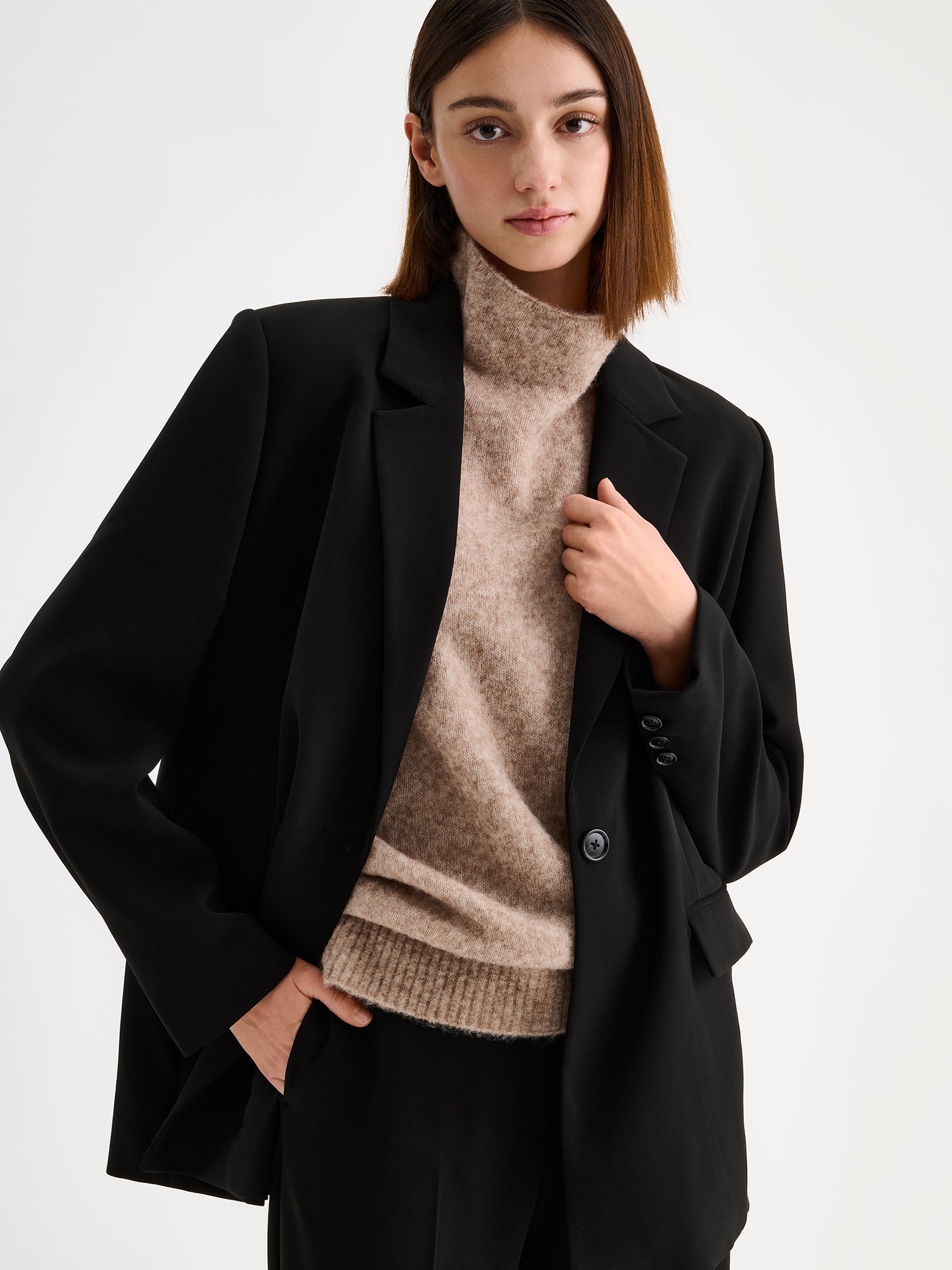 A stylish women's Eeva blazer in black with a relaxed yet structured silhouette, on a model with matching wide legged trousers and a beige knitted vest.
