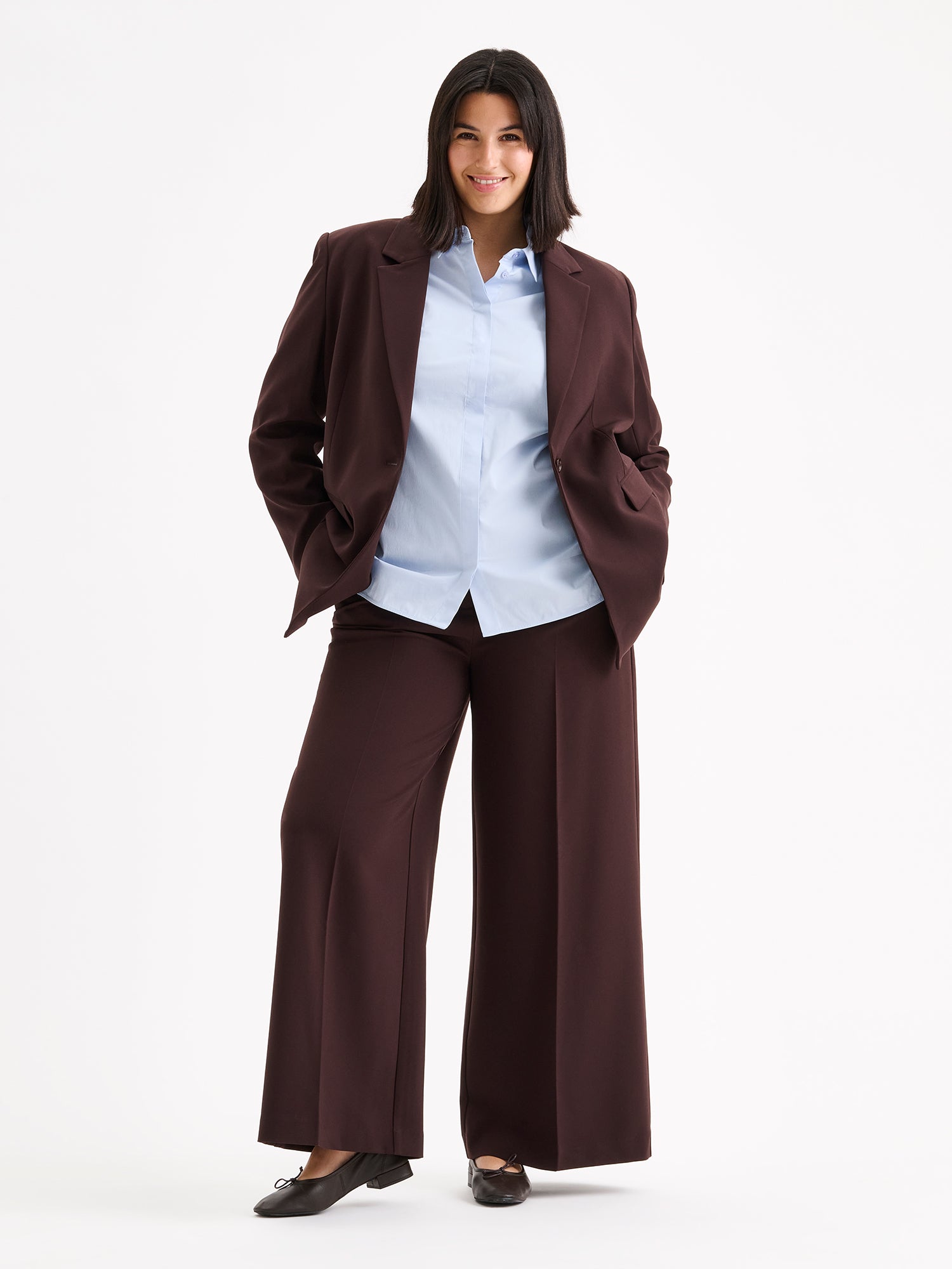A stylish women's Eeva blazer in a rich brown shade with a relaxed yet structured silhouette, on a model with a light blue shirt and ballerinas