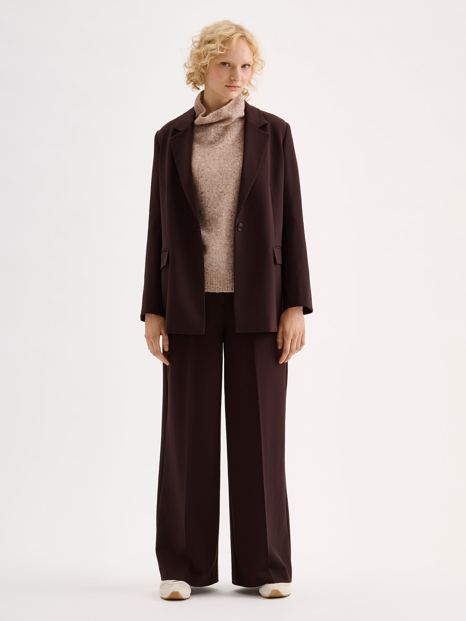 A stylish women's Eeva blazer in a rich brown shade with a relaxed yet structured silhouette, on a model with a beige knitted vest and sneakers