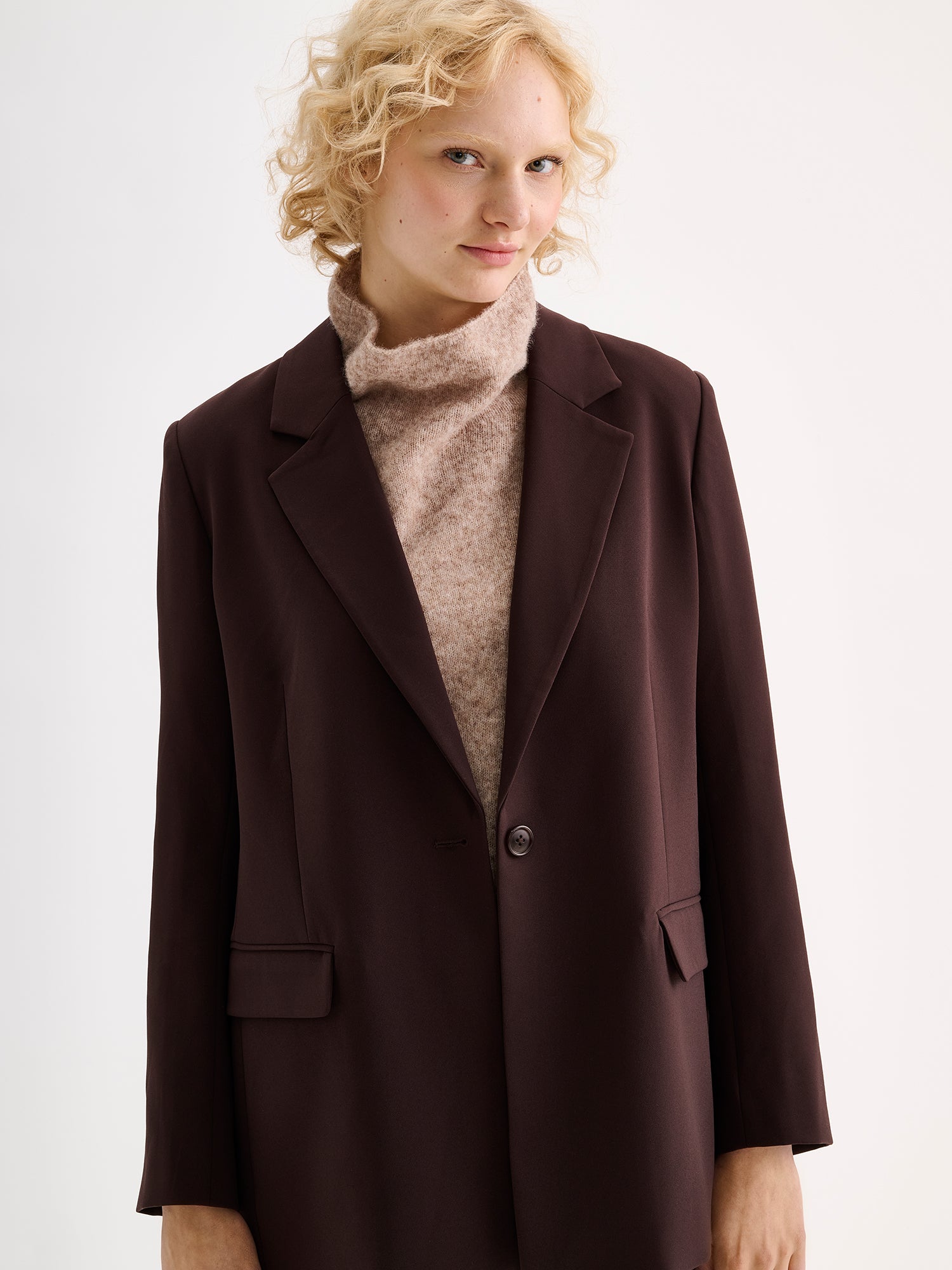 A stylish women's Eeva blazer in a rich brown shade with a relaxed yet structured silhouette, on a model with a beige knitted vest