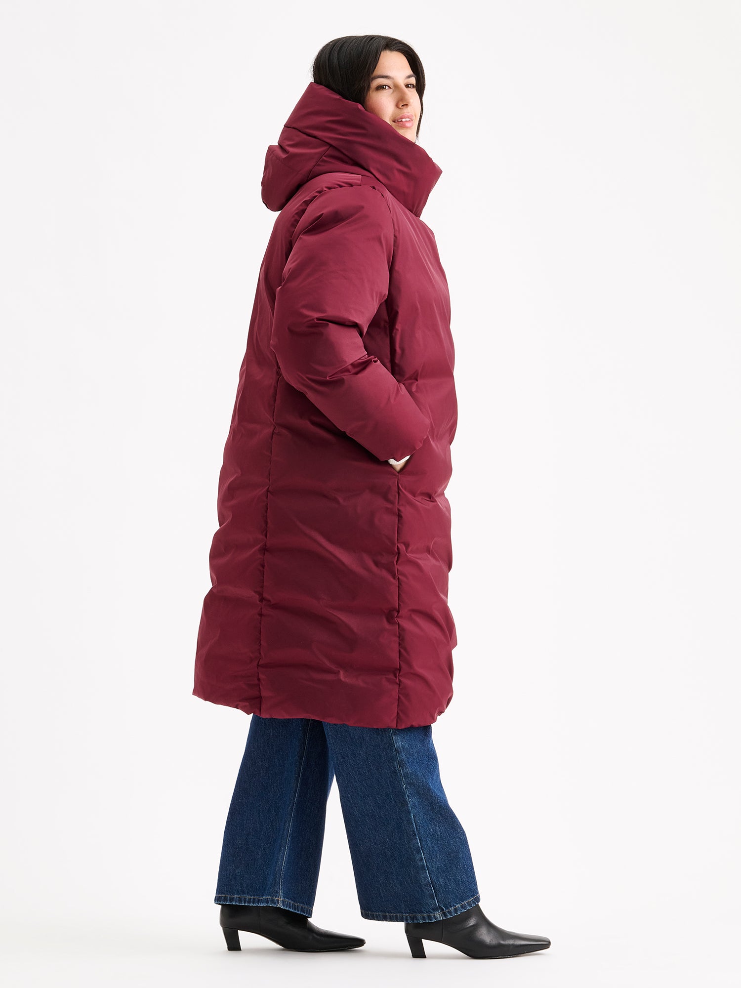 Warm and wonderfully voluminous, the long Inari puffer coat in winered on a model with jeans, side view.