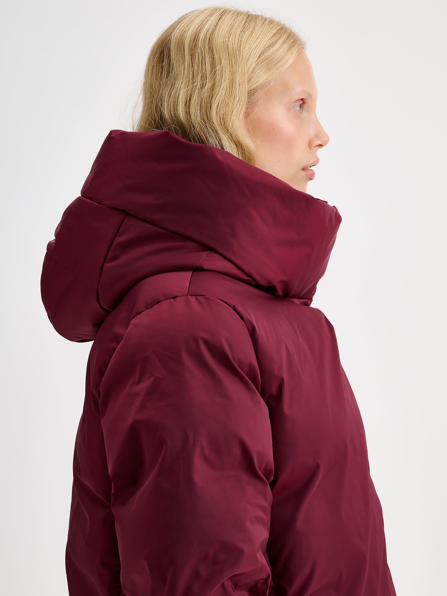 Warm and wonderfully voluminous, the long Inari puffer coat in winered on a model close-up from the side.